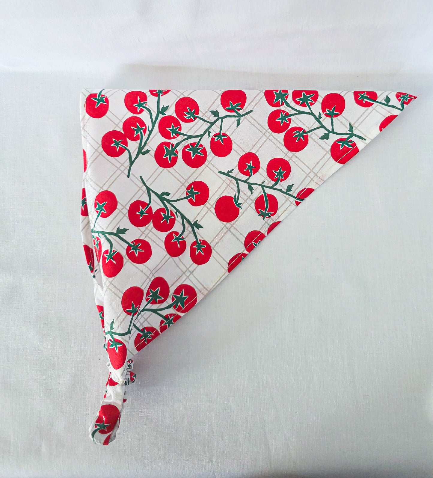 A triangular bandana made of white fabric with a red tomato print and green stems with an elastic band.