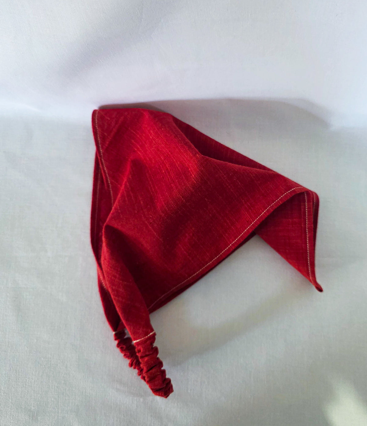 A folded red fabric bandana with a gathered elasticized band, displayed on a white fabric background.