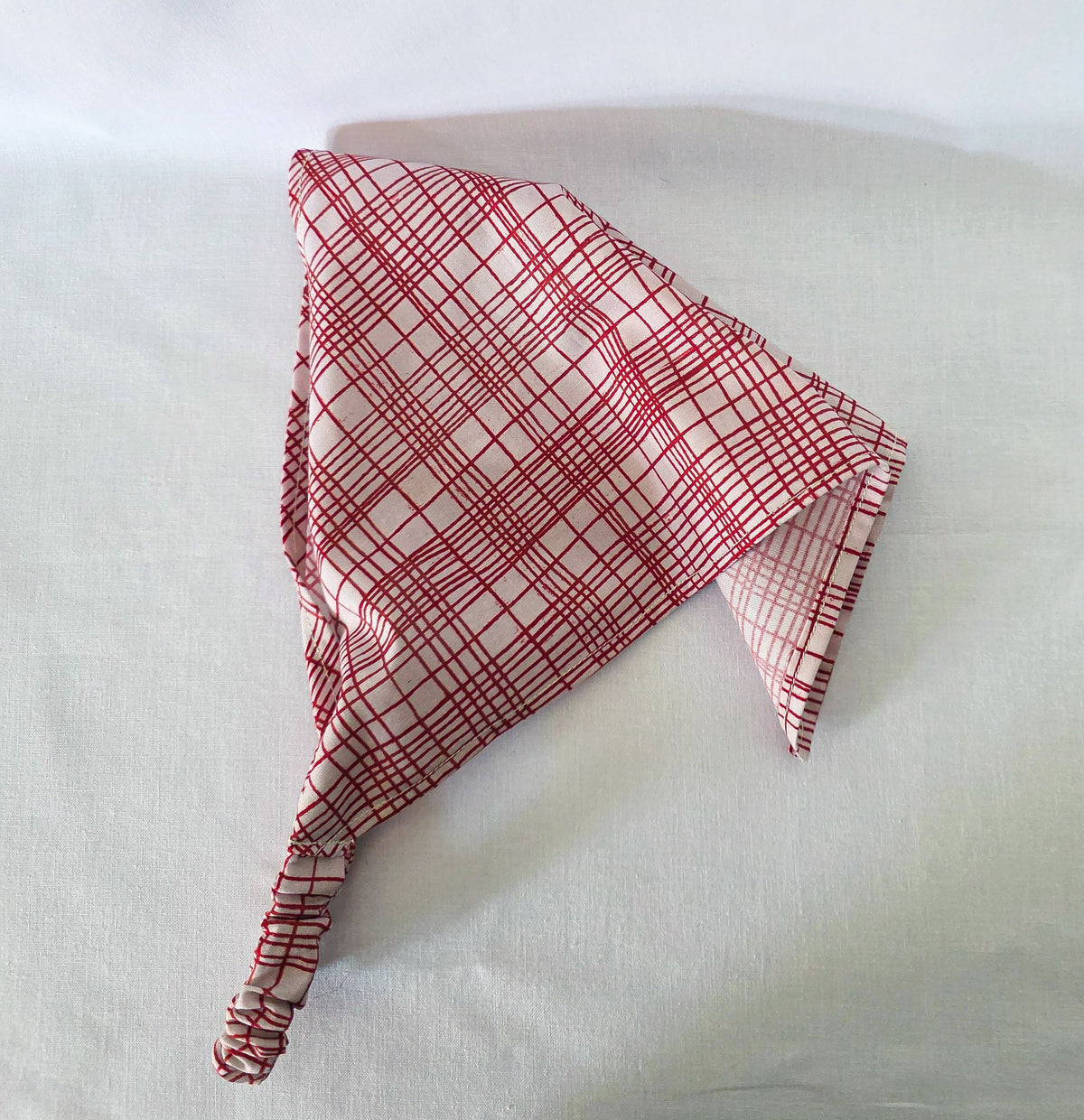 A red and white plaid hair bandana with an elastic band, laid flat on a white fabric background.