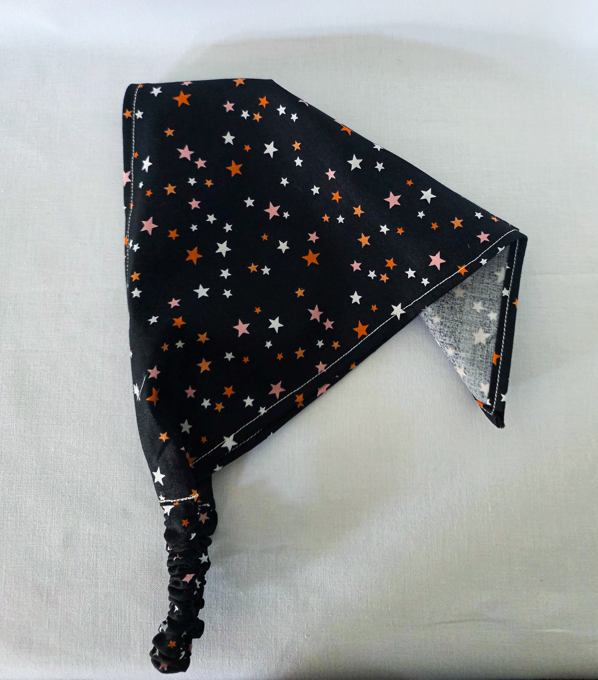 A triangular hair bandana with an elastic neck loop, featuring a black background printed with small stars in orange, pink, and white. The bandana is laid flat on a white fabric background.