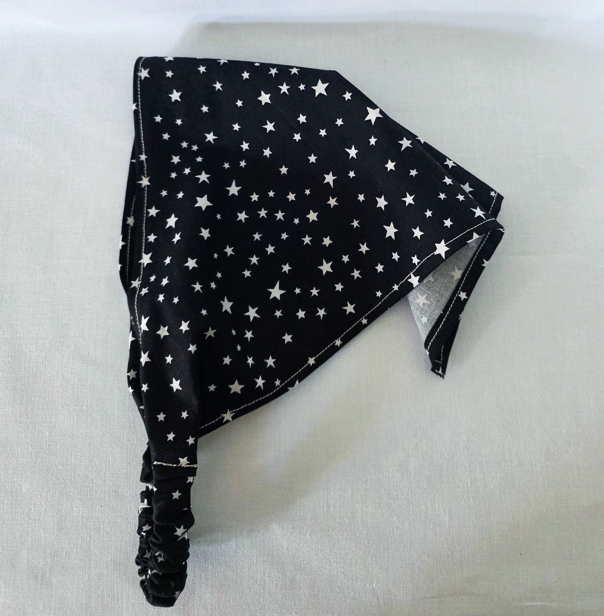 A black hair bandana with a white star pattern, folded and displayed on a light-colored fabric background.