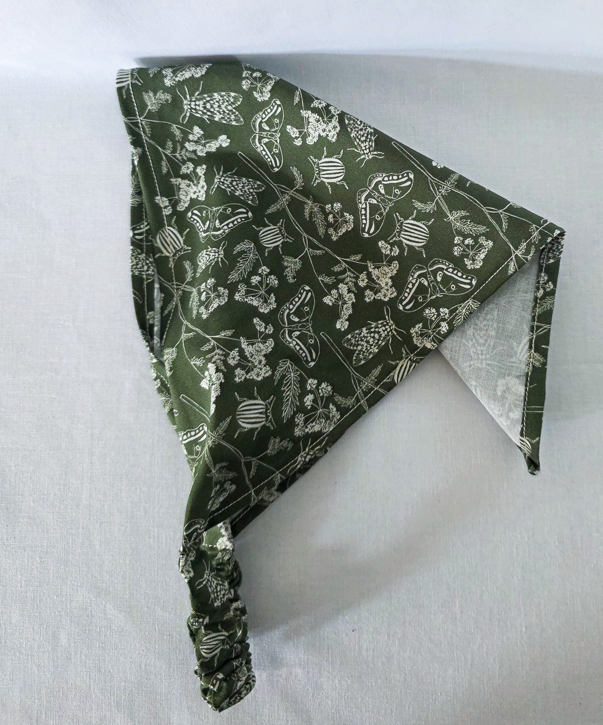 A green elastic hair bandana with a white floral and bug print on it. Laid on its side showing it drape, on a white fabric background.