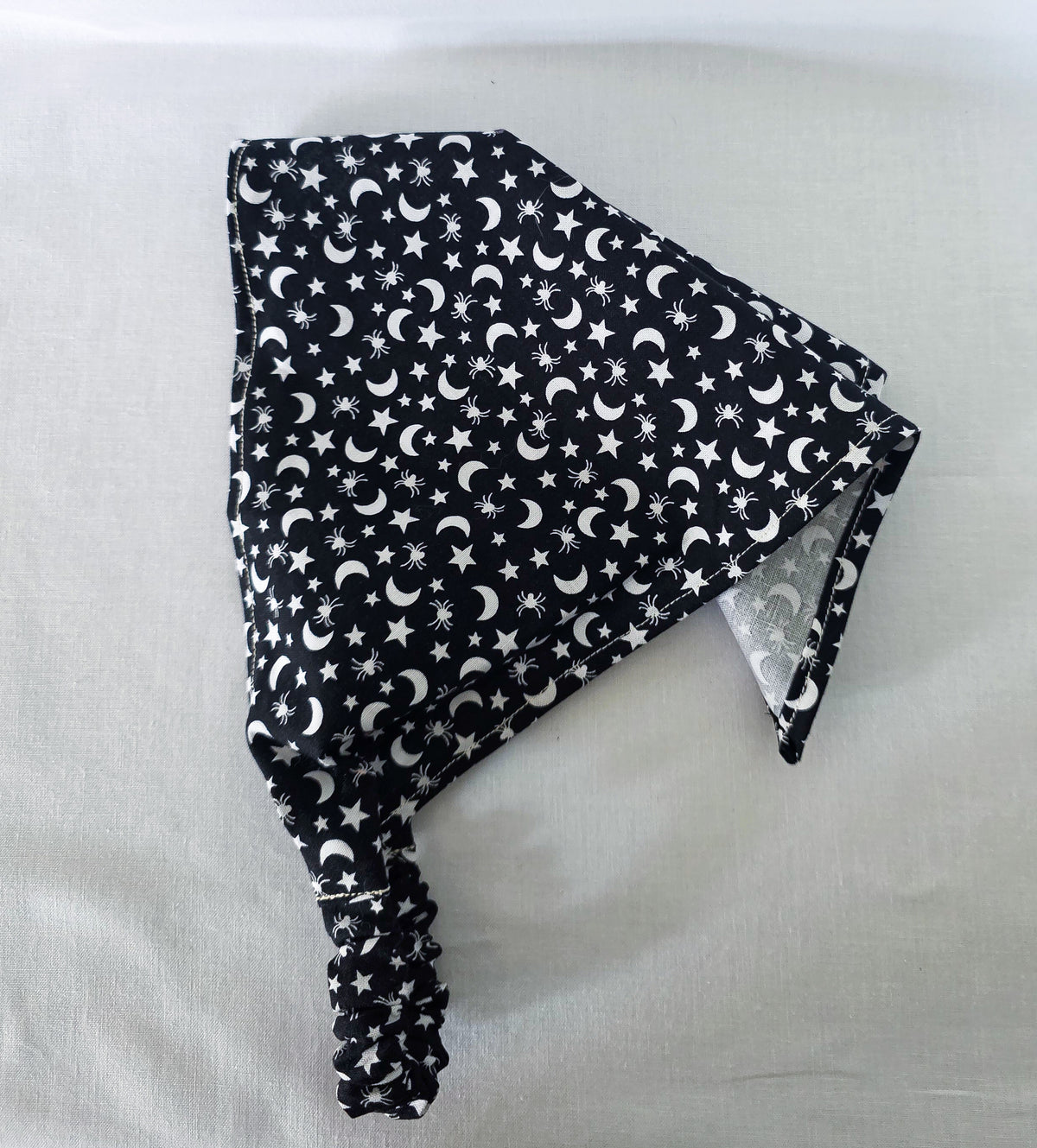 A black fabric elastic hair bandana with a white celestial pattern of crescent moons, stars, and spiders draped on its side on a white fabric background.