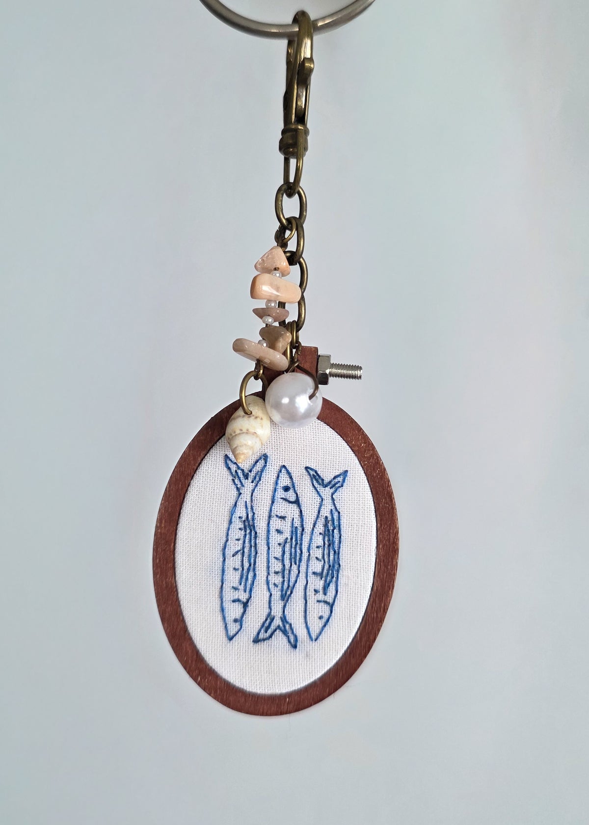 hand embroidered keychain featuring embroidered outlines of three sardines, done in blue thread, in an oval wooden hoop. It has a shell and pearl charm. Hanging against a light background.