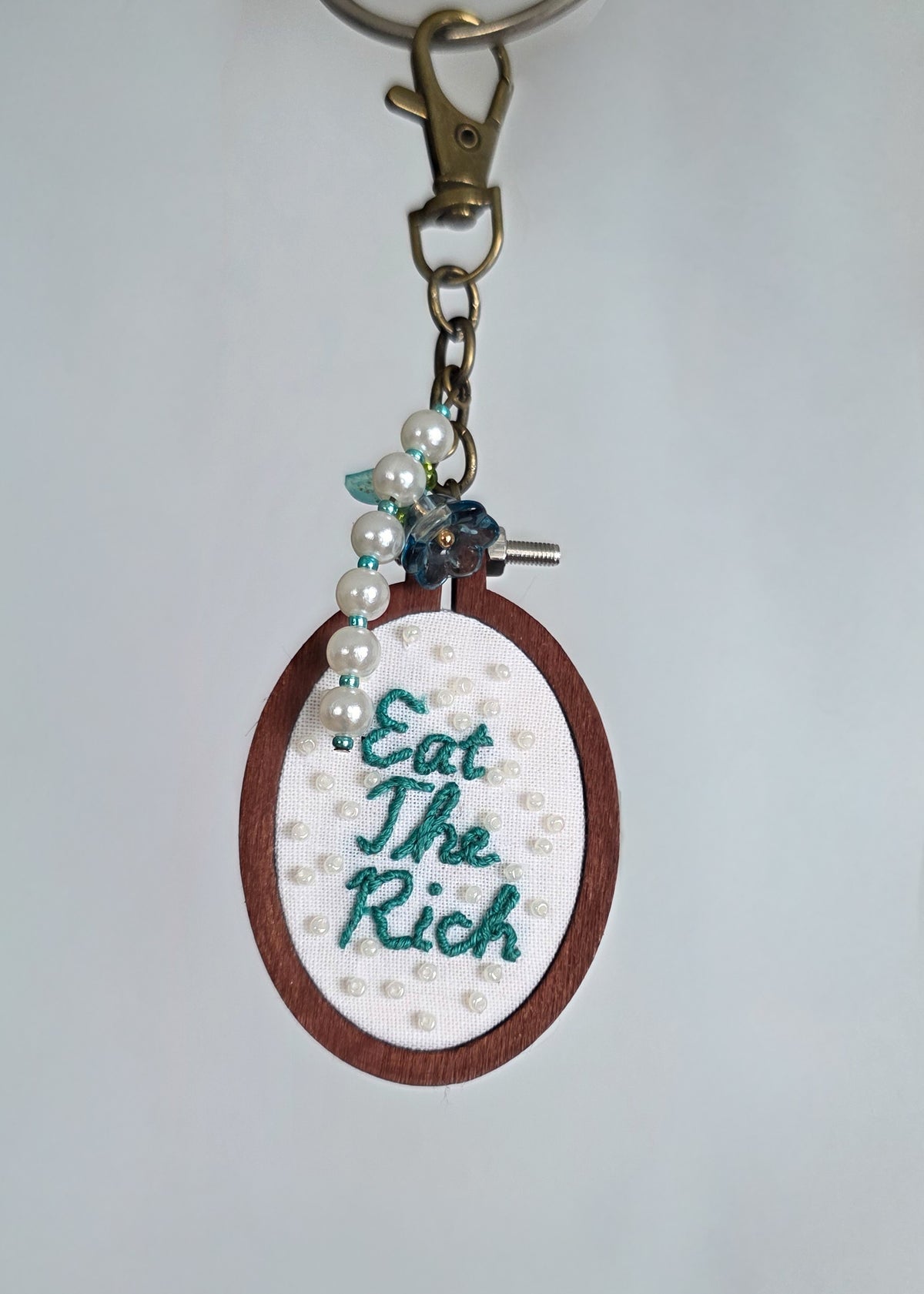 Hand embroidered keychain featuring the phrase "Eat The Rich". it has embroidered white beads on it, and has a floral and pearl charm. It is hanging against a grey background.