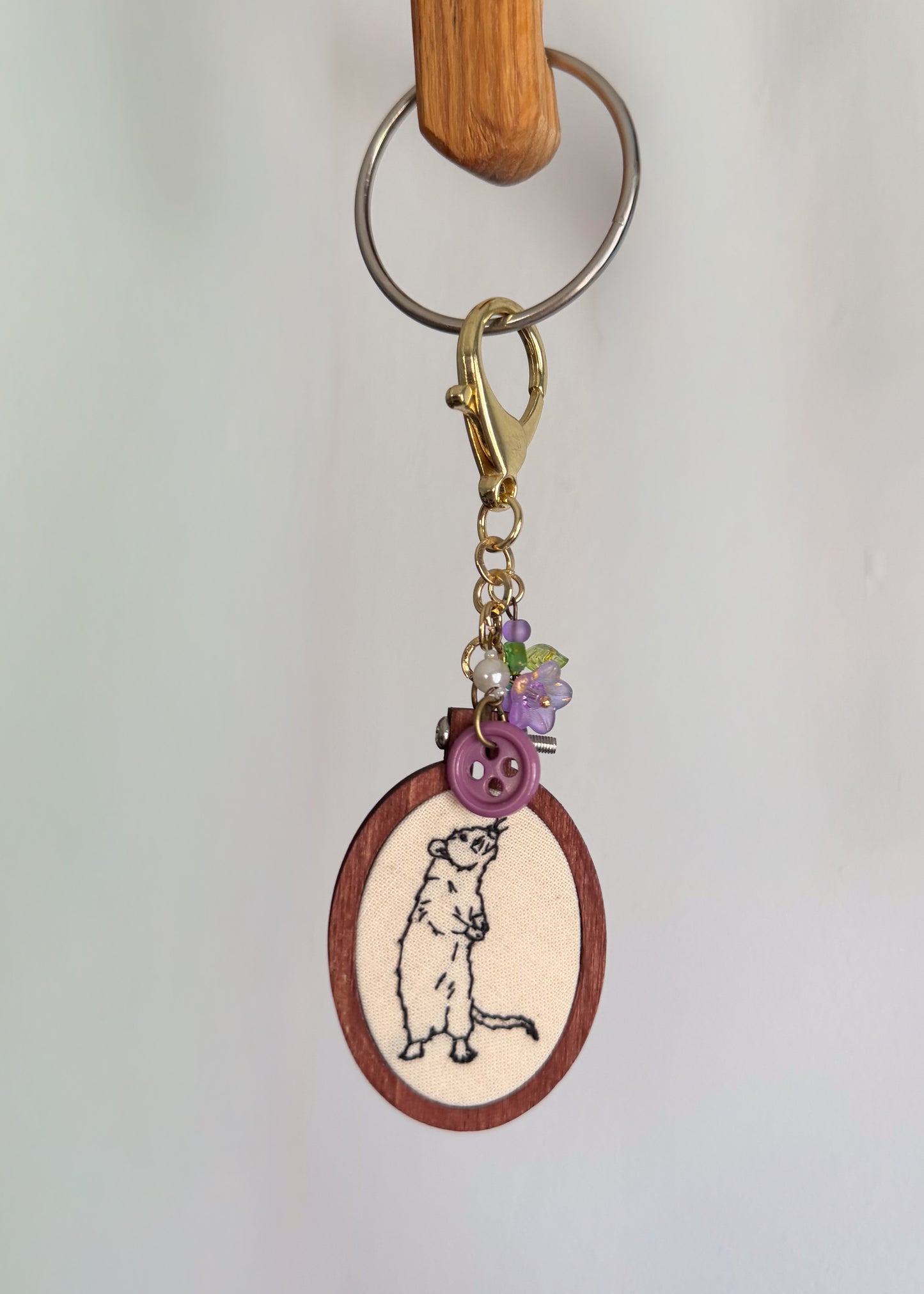 A handmade keychain featuring a wooden oval charm with an embroidered mouse design, adorned with a purple button, purple flower charms, and a gold clasp, hanging from a metal ring on a wooden peg against a white wall.