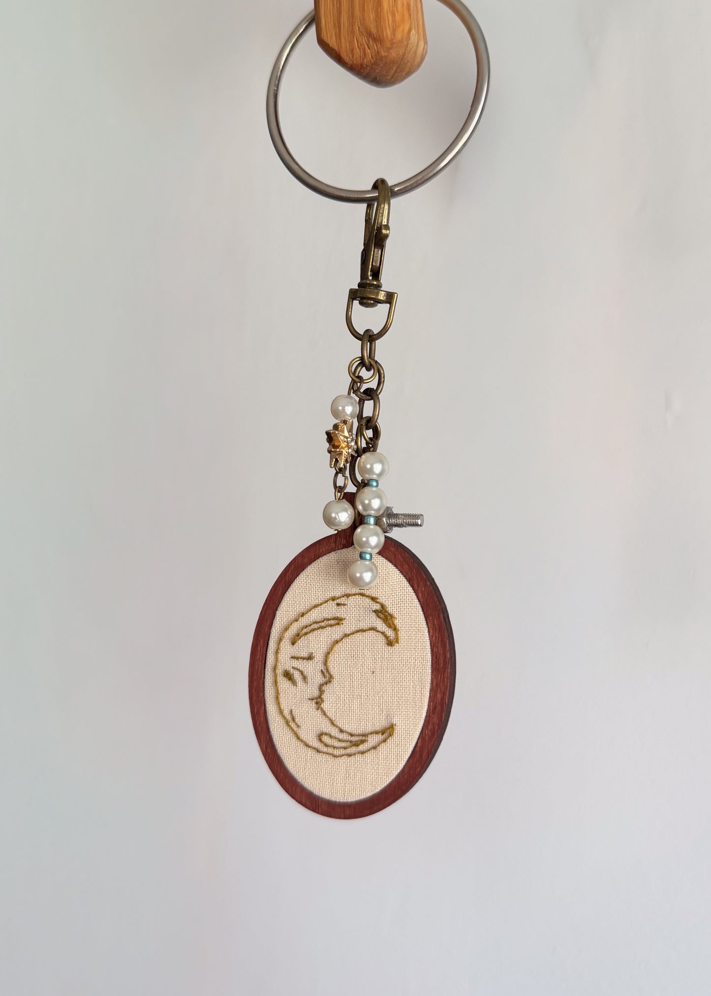 A handmade keychain featuring a circular wooden medallion with a hand embroidered crescent moon design, hanging from a metal split ring and clasp.