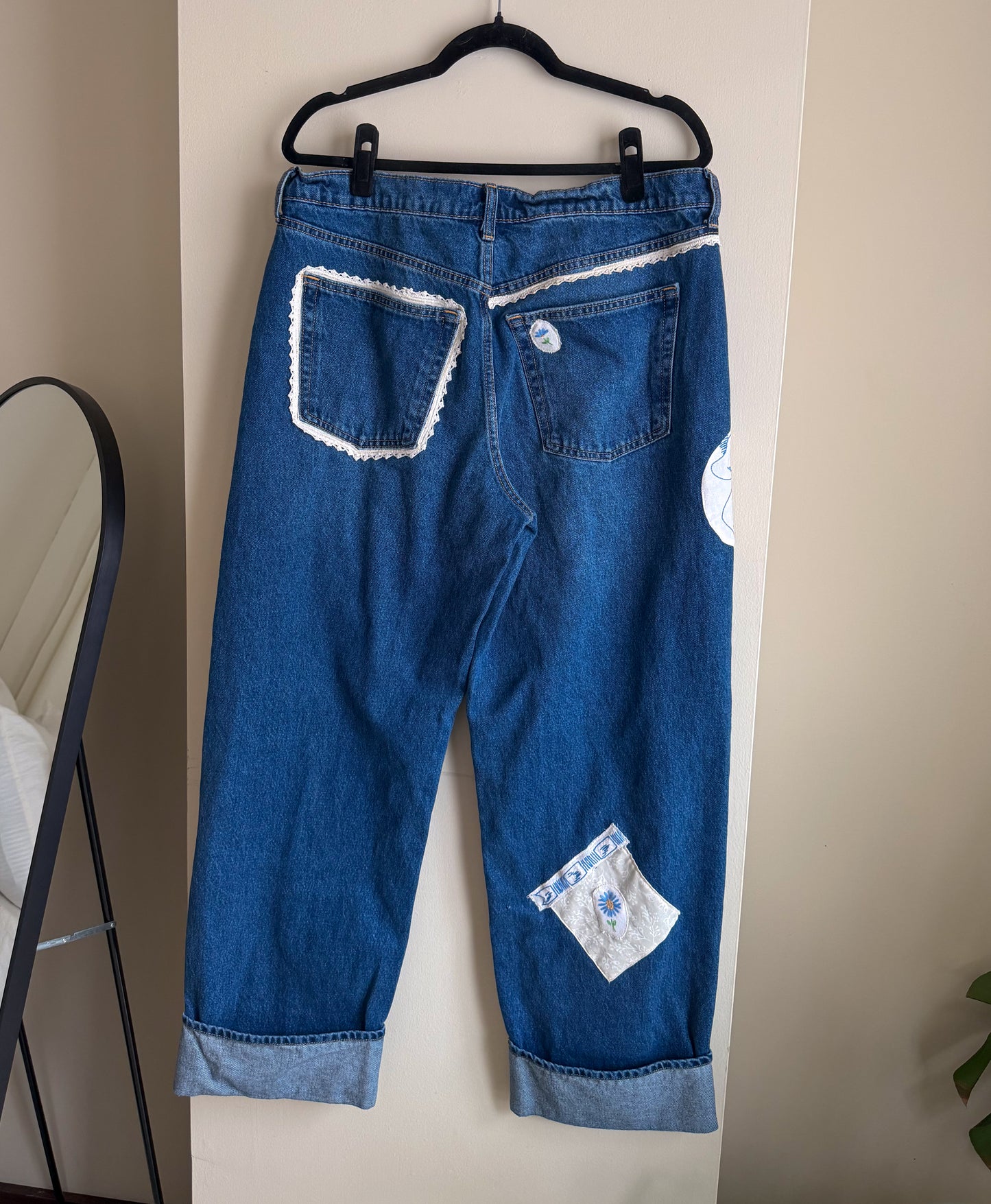 Blue jeans with white patches and lace on a hanger against a beige wall.