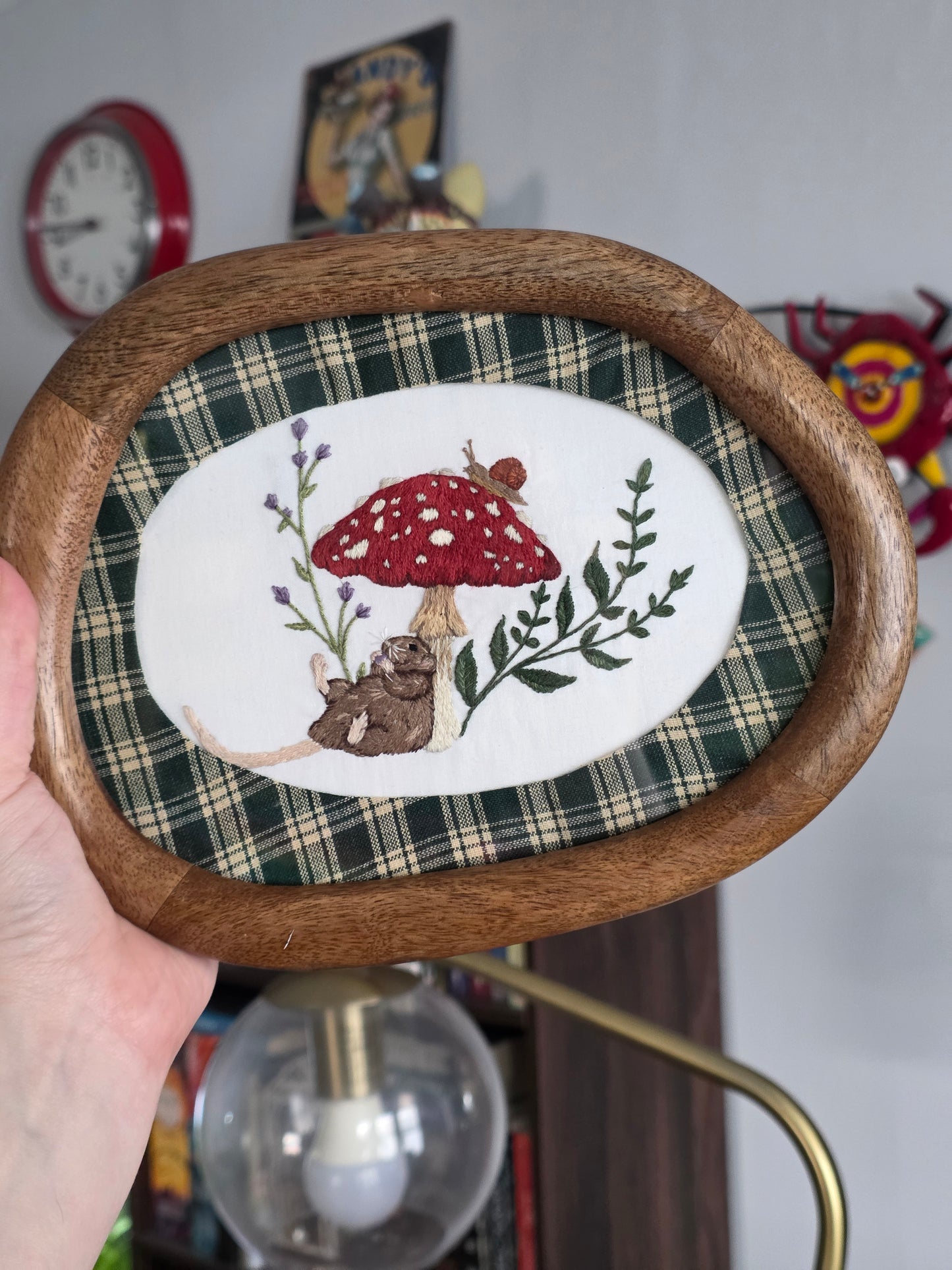 A hand holds an oval wooden-framed piece of embroidered wall art. The design features a red mushroom with white spots, green leaves, and a brown rat on a white background with a green plaid border. This is the original artwork of the print.