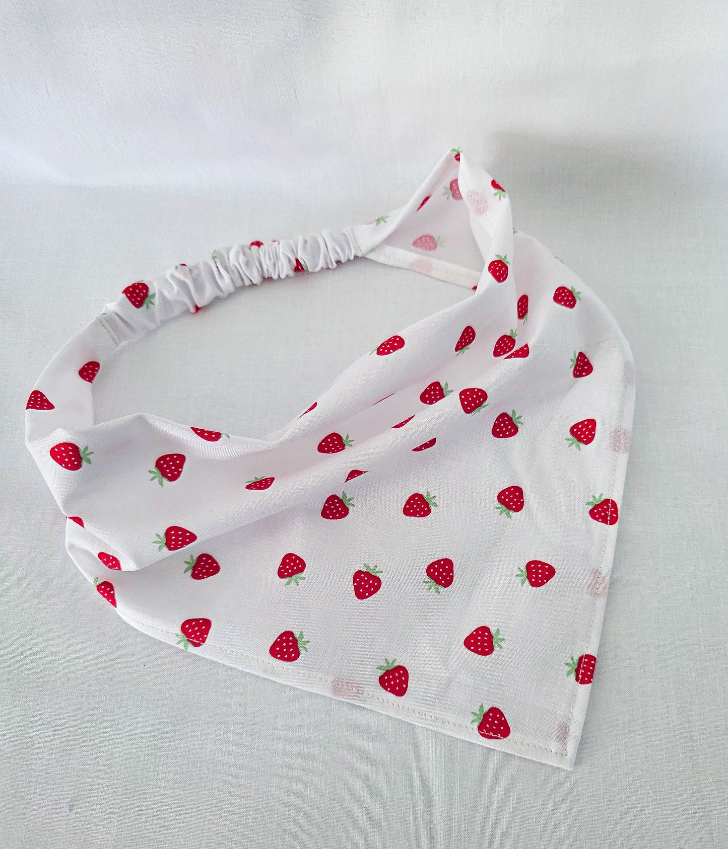 A white triangular hair bandana with a repeating red strawberry pattern, featuring a gathered elastic head strap, laid flat on a white fabric background.