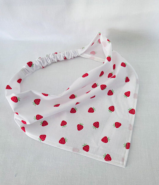 A white triangular hair bandana with a repeating red strawberry pattern, featuring a gathered elastic head strap, laid flat on a white fabric background.