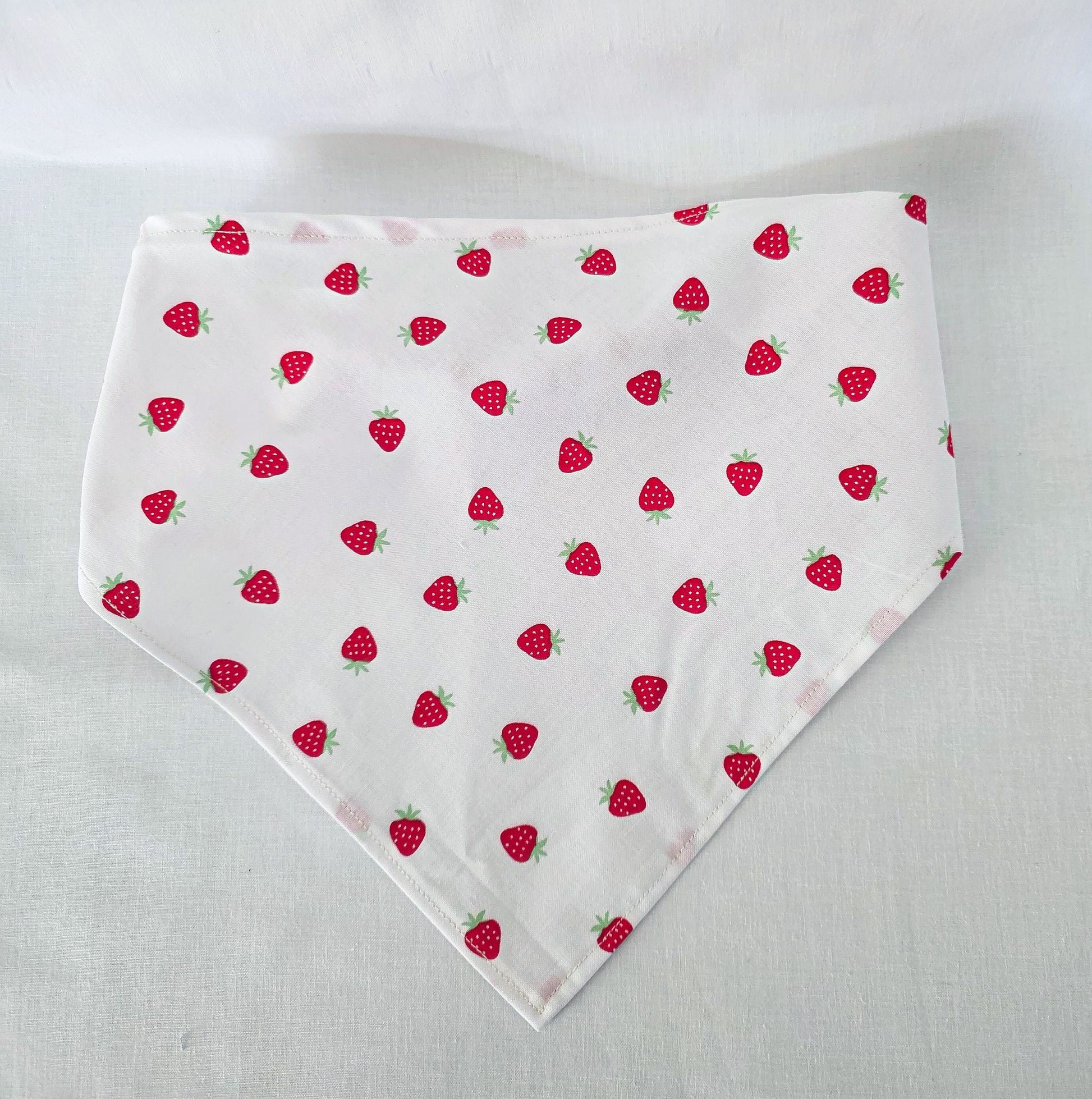 A white triangular bandana or bib-style fabric accessory with a repeating red strawberry print and green leafy tops, laid flat on a light fabric background.