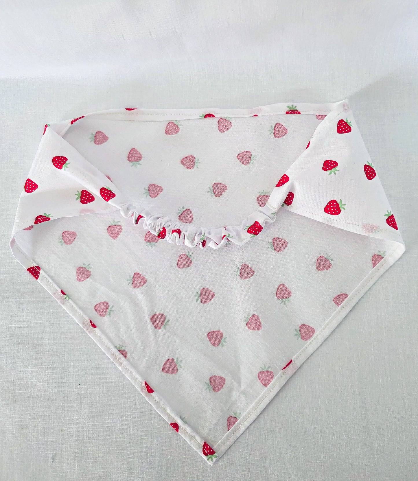 A white triangular bandana with a repeating pattern of red strawberries, laid flat on a light fabric background.