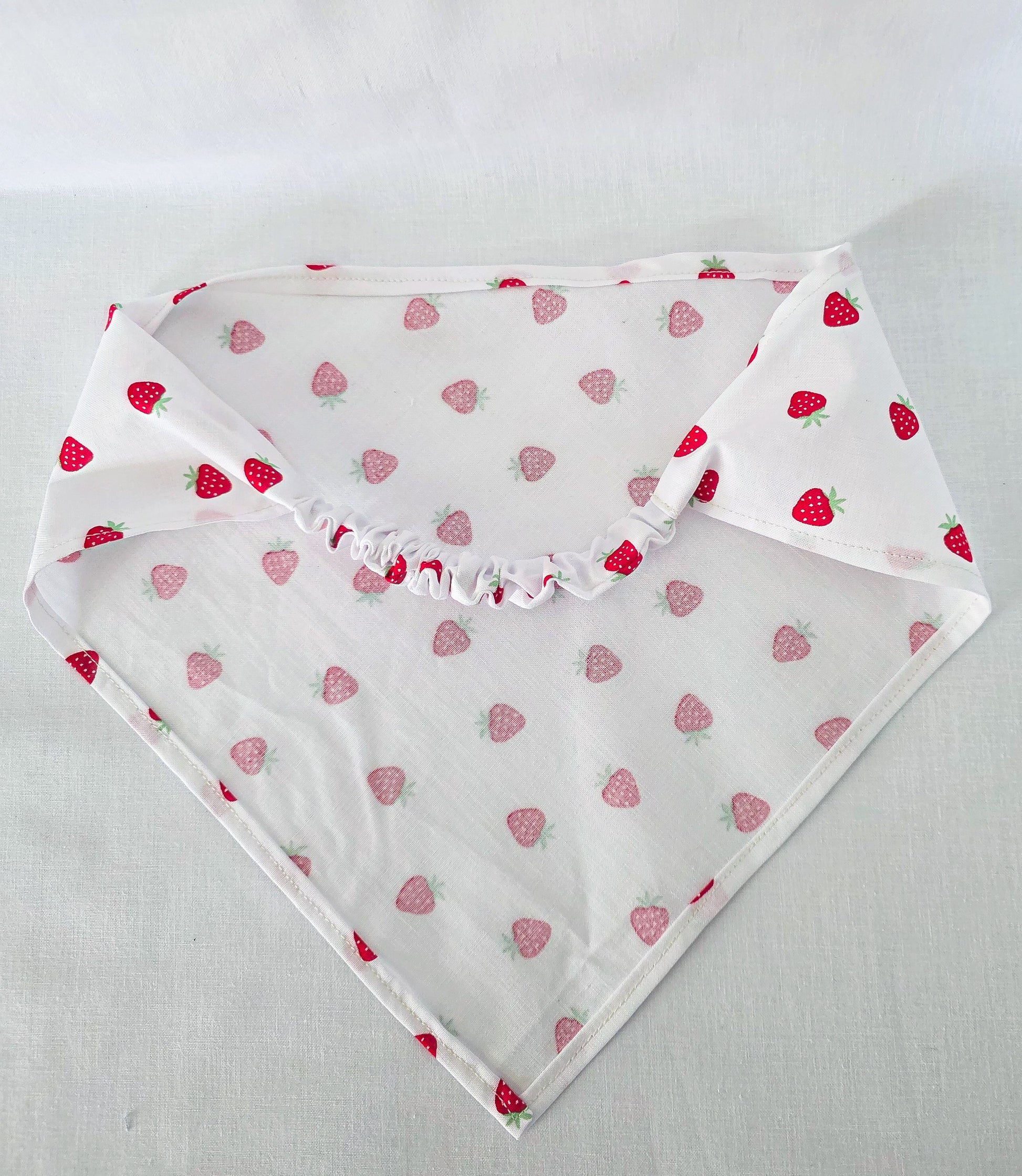 A white triangular bandana with a repeating pattern of red strawberries, laid flat on a light fabric background.