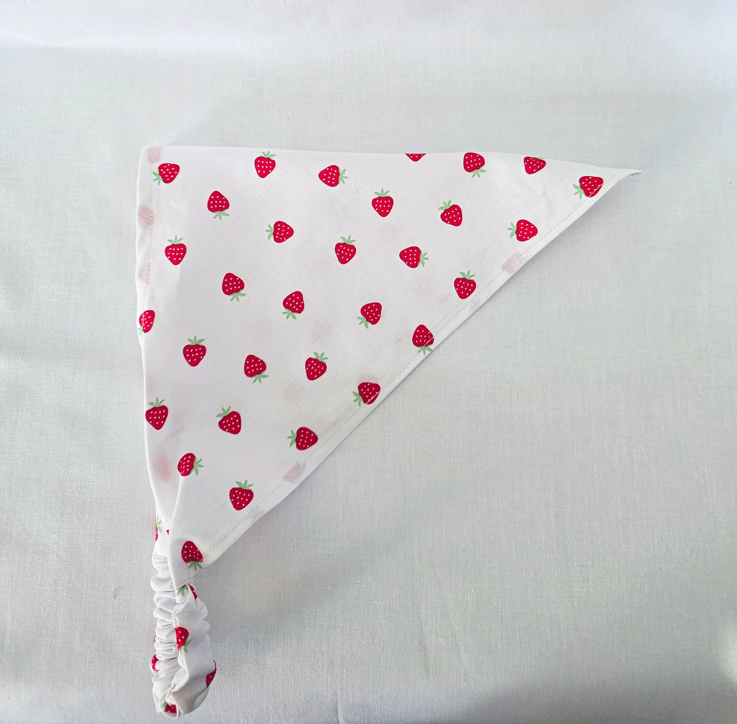 A white triangular bandana  with a repeating red strawberry pattern, featuring a gathered elasticized bottom corner, laid flat on a white fabric background.