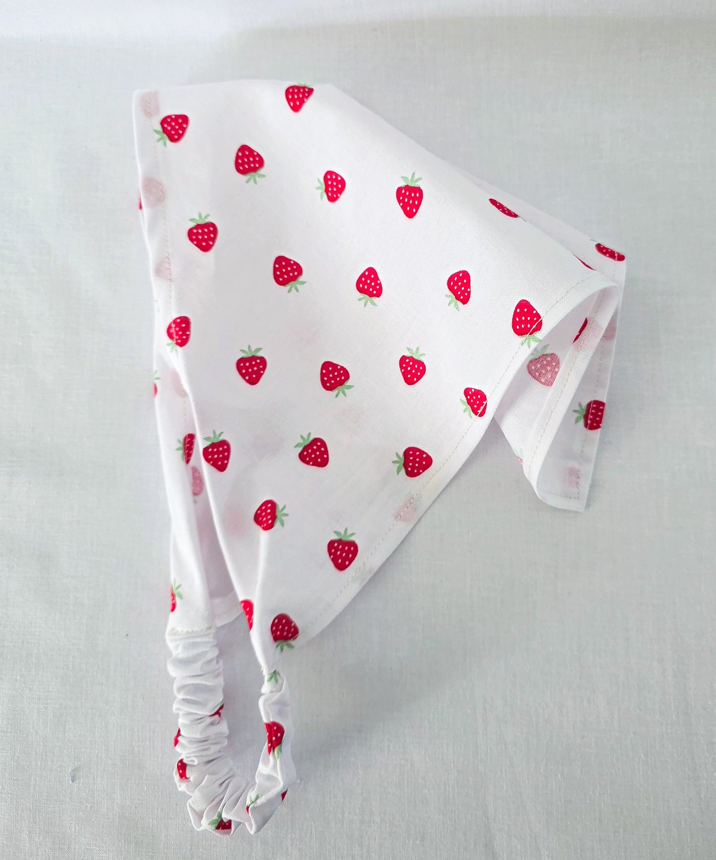 A white hair bandana with a red strawberry pattern, featuring a gathered elasticized edge, laid flat on a light fabric background.
