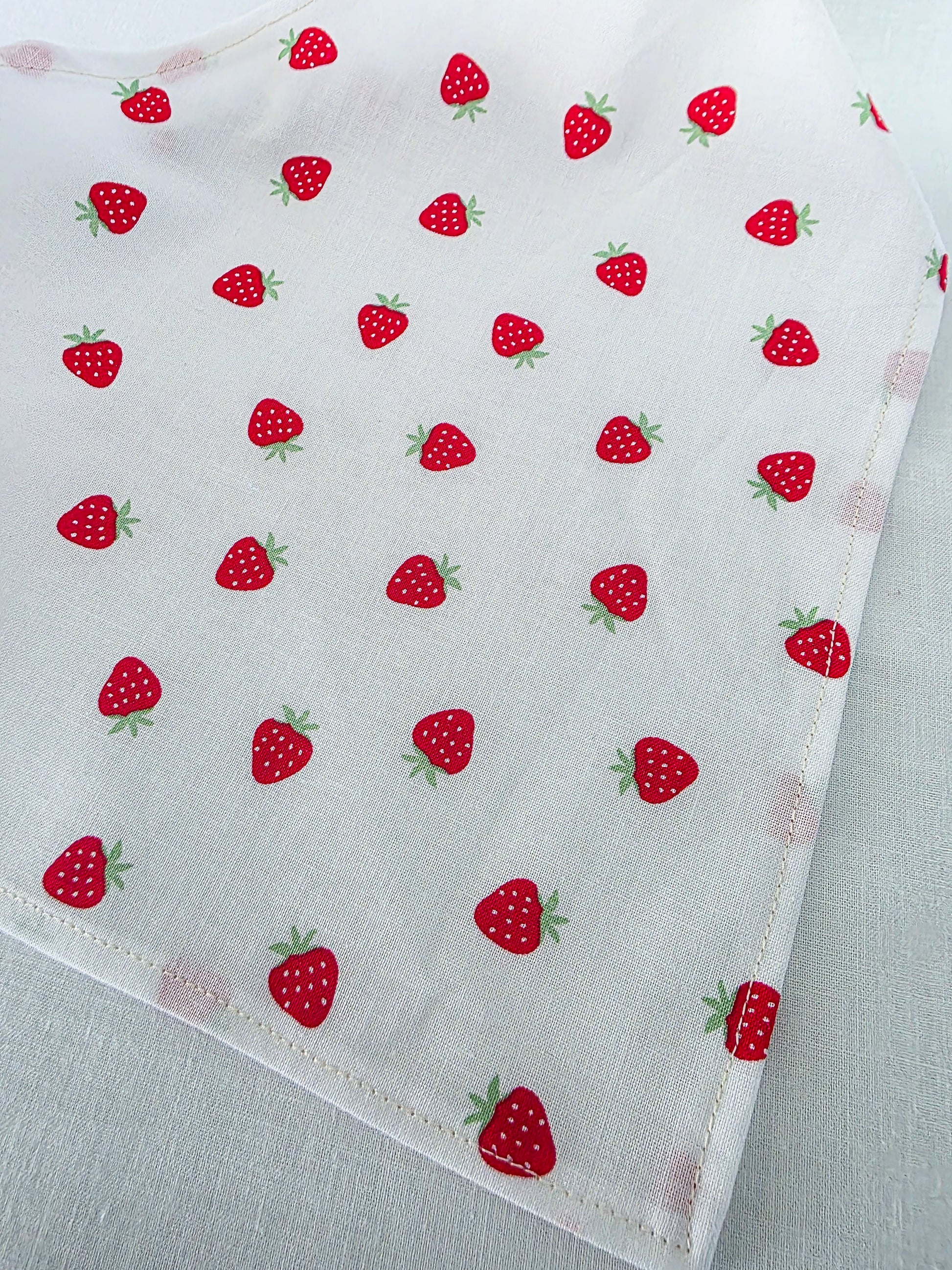 White fabric with a repeating pattern of red strawberries with green leaves, laid flat on a light fabric background.