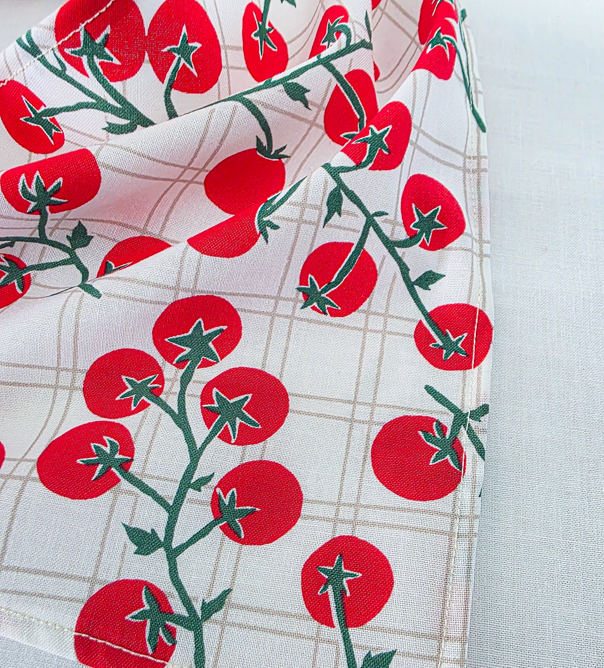 A close-up of a white fabric with a red tomato print and a light beige plaid pattern, draped to show the texture and design.