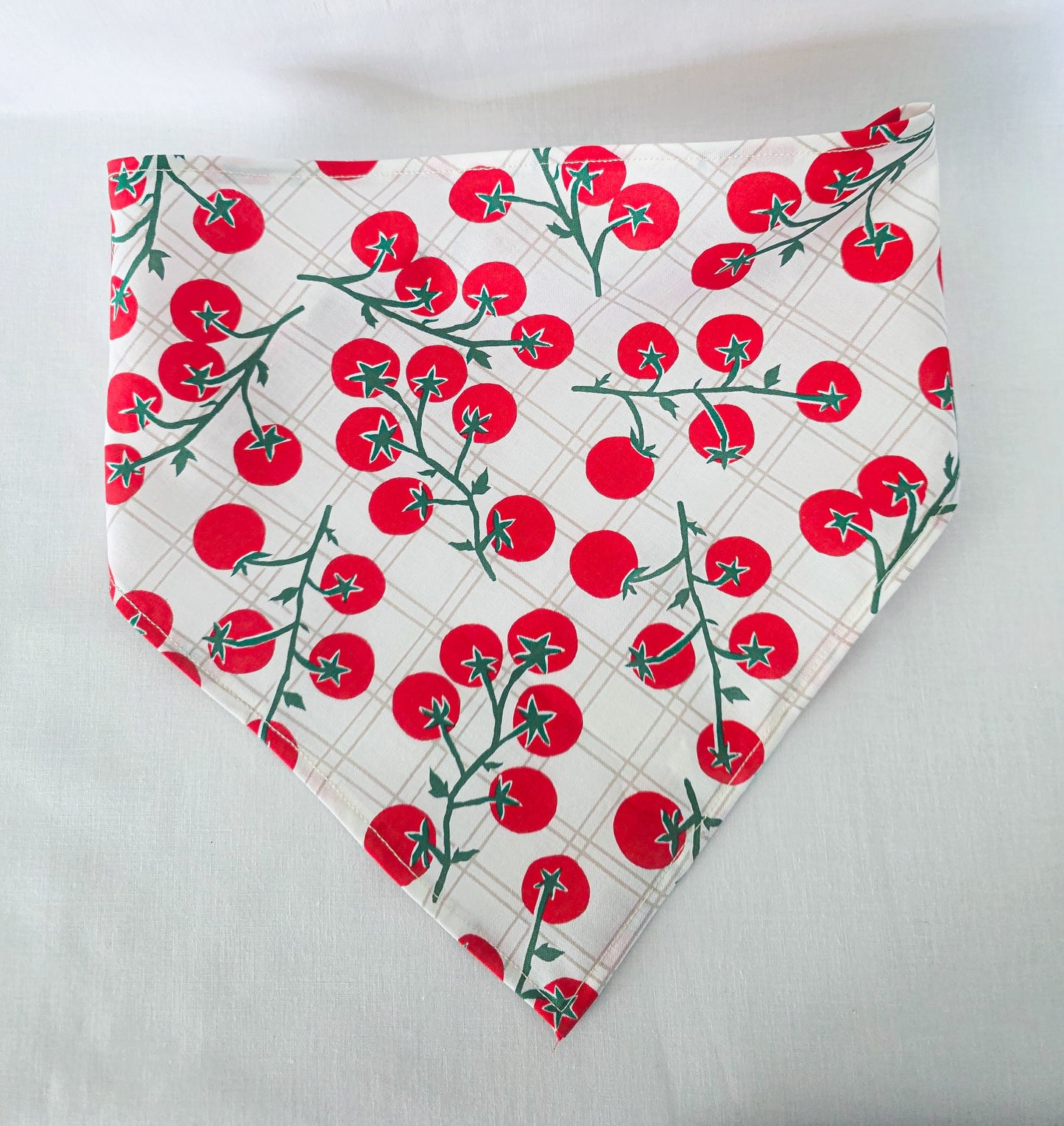 A triangular hair bandana with a white background, featuring a repeating pattern of red cherry tomatoes on green vines over a subtle beige plaid grid.