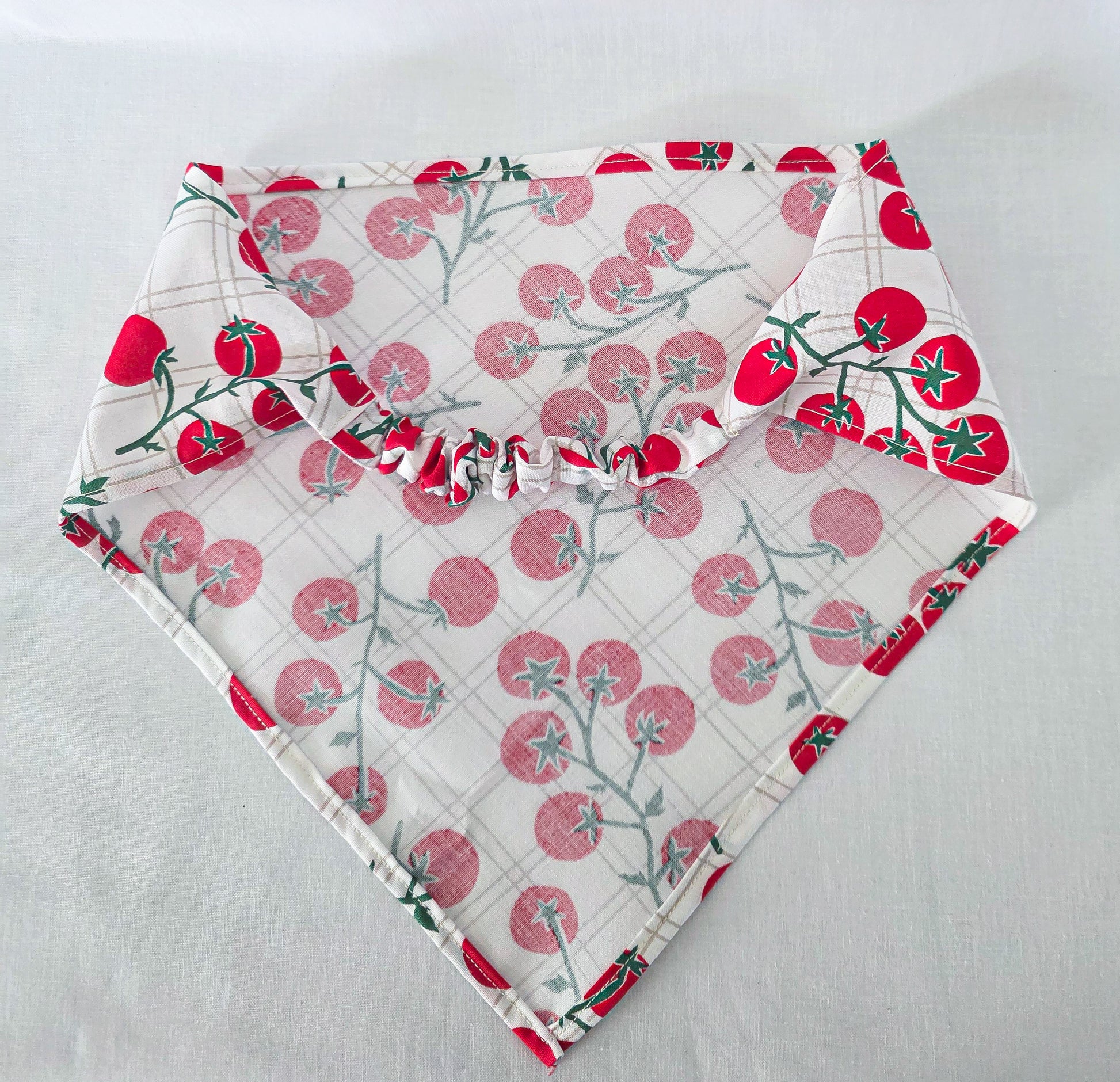 A hair bandana laid flat on a white fabric background, featuring a red tomato print with green stems and a subtle gray plaid pattern.