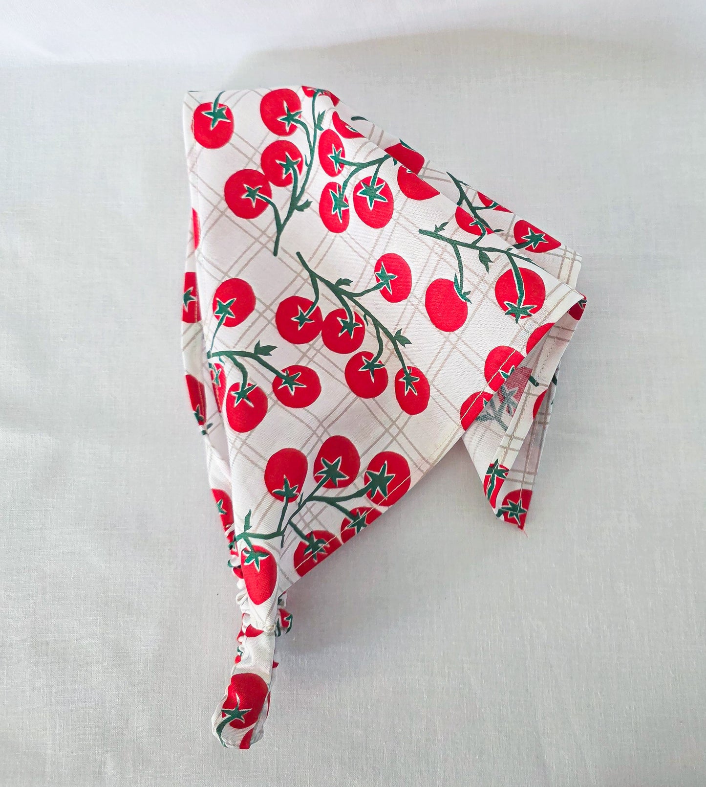A triangular fabric hair bandana featuring a red tomato print on a white background with a subtle plaid pattern. The fabric is laid flat on a light-colored textured surface.