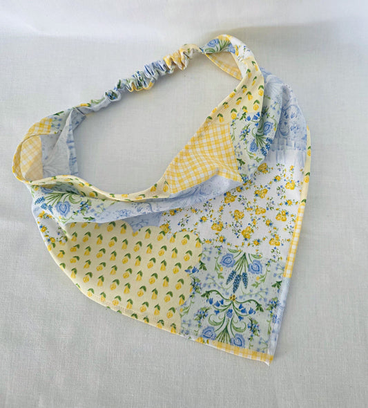 A triangular hair bandana laid flat on a light beige fabric background. The bandana features a multi-patterned design with yellow and white gingham, yellow floral prints, and blue floral motifs.
