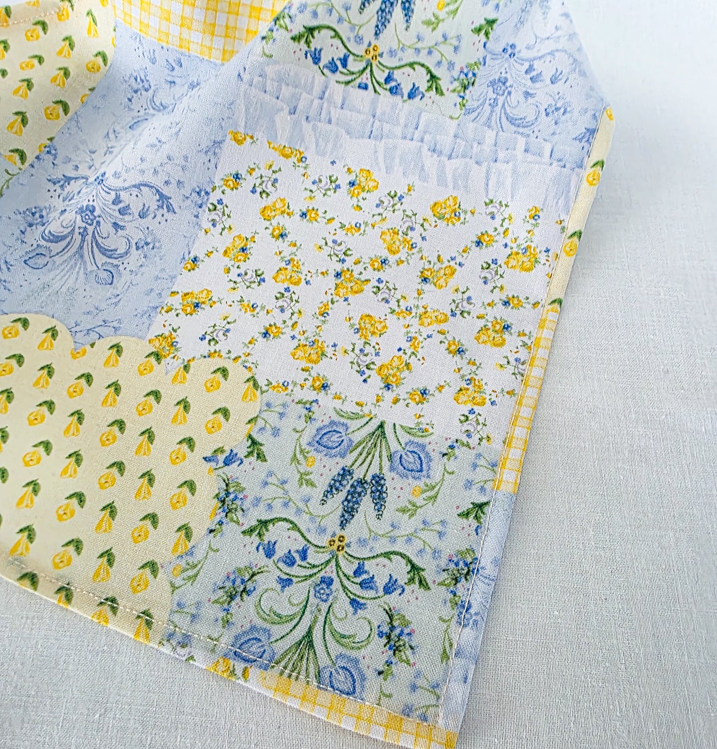A close-up of a hair bandana featuring a faux patchwork design of floral and fruit prints including blue flowers, yellow blossoms, on a white and light blue background.