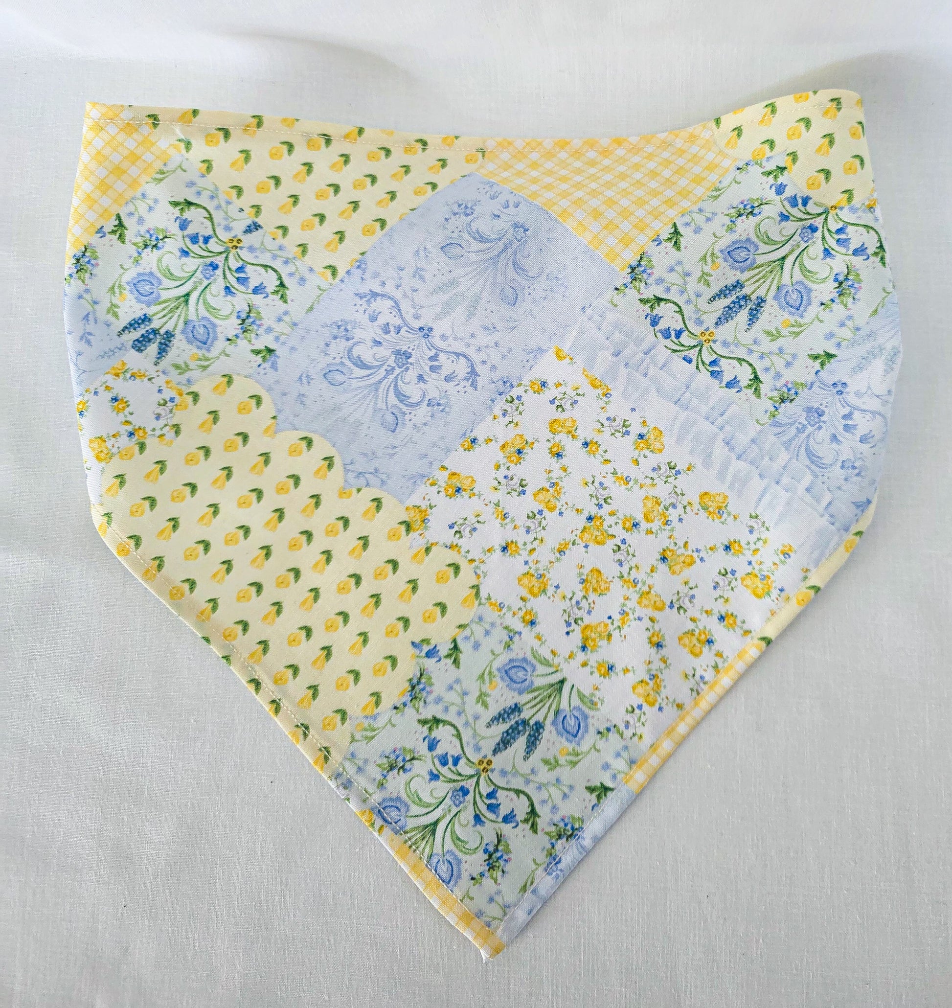 A handmade triangular hair bandana with a patchwork design featuring yellow prints, blue floral patterns, and a light blue gingham check, laid flat on a white fabric background.
