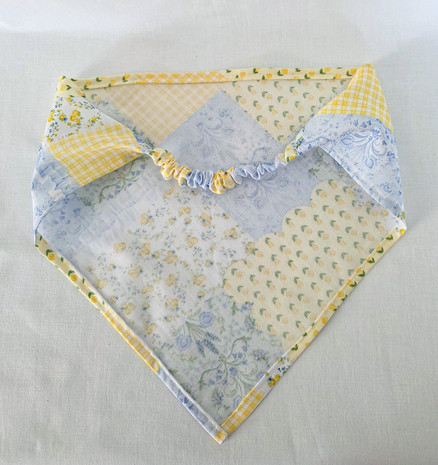 A handmade faux patchwork hair bandana  laid flat on a light fabric background. The bib features a variety of patterns including yellow gingham, light blue paisley, and floral prints in yellow and blue, with an elastic band.