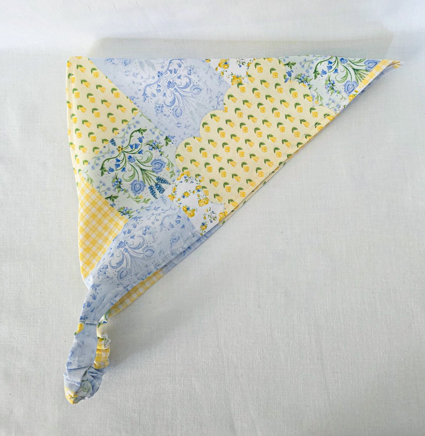 A triangular hair bandana with a faux patchwork design featuring yellow and white patterns, blue floral prints, and a yellow-and-white gingham section and displayed on a light gray fabric background.