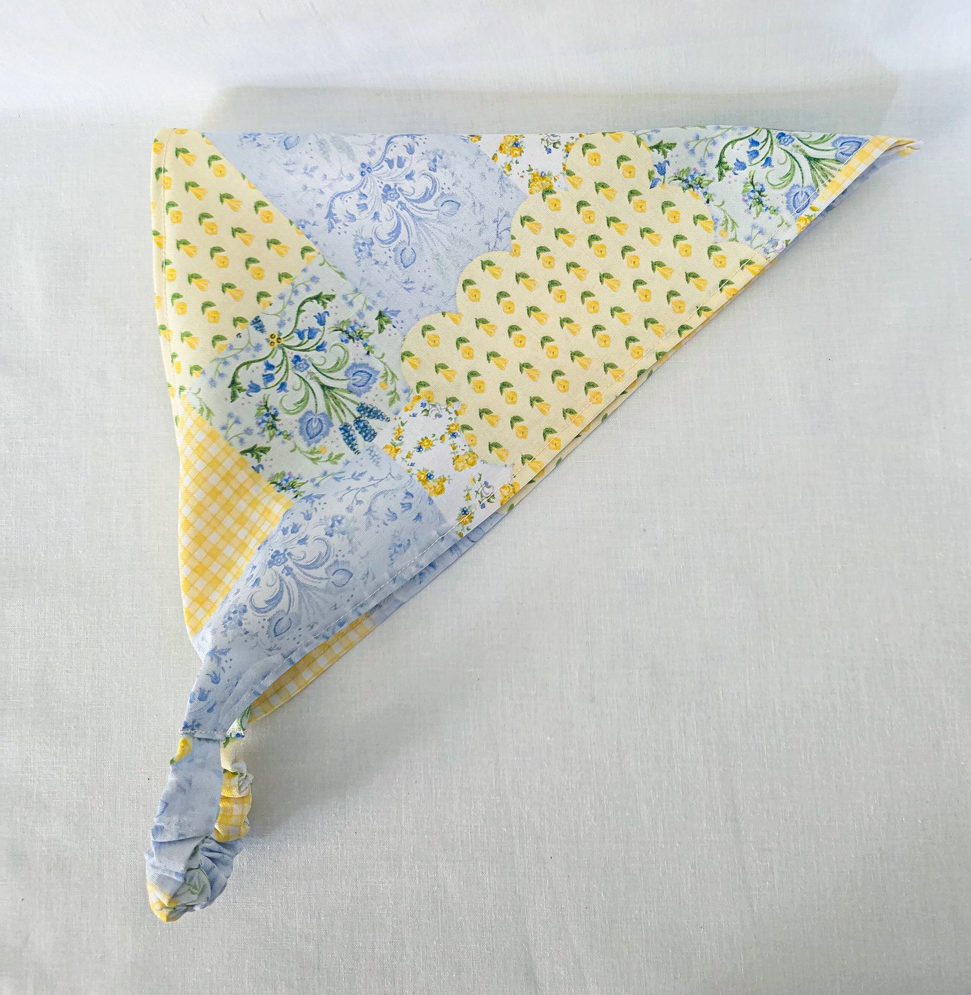 A triangular hair bandana with a faux patchwork design featuring yellow and white patterns, blue floral prints, and a yellow-and-white gingham section and displayed on a light gray fabric background.
