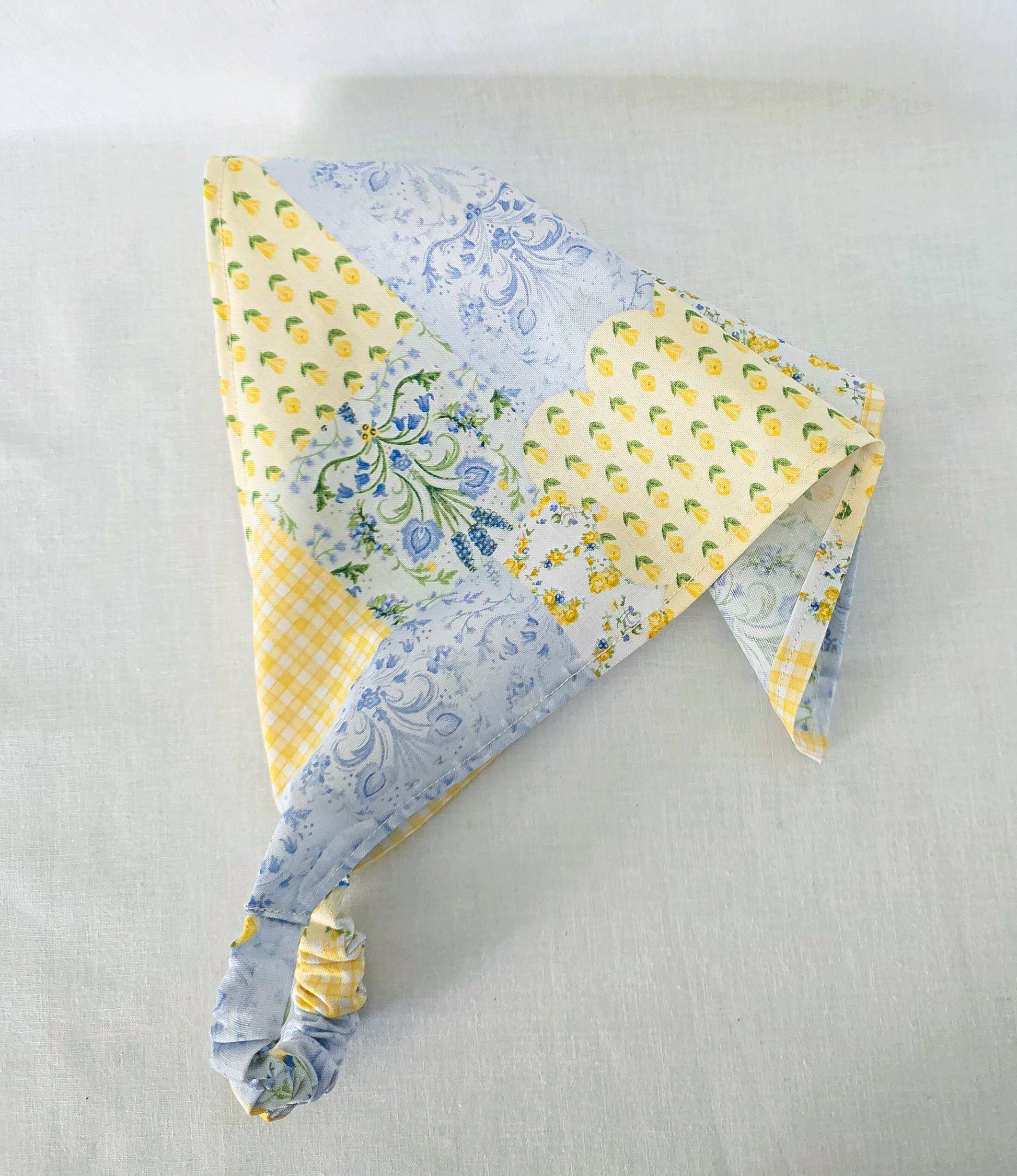 A triangular hair bandana a made from a faux patchwork of light blue floral, yellow l prints, and yellow gingham fabrics, and an elastic band. Displayed on a light beige fabric background.