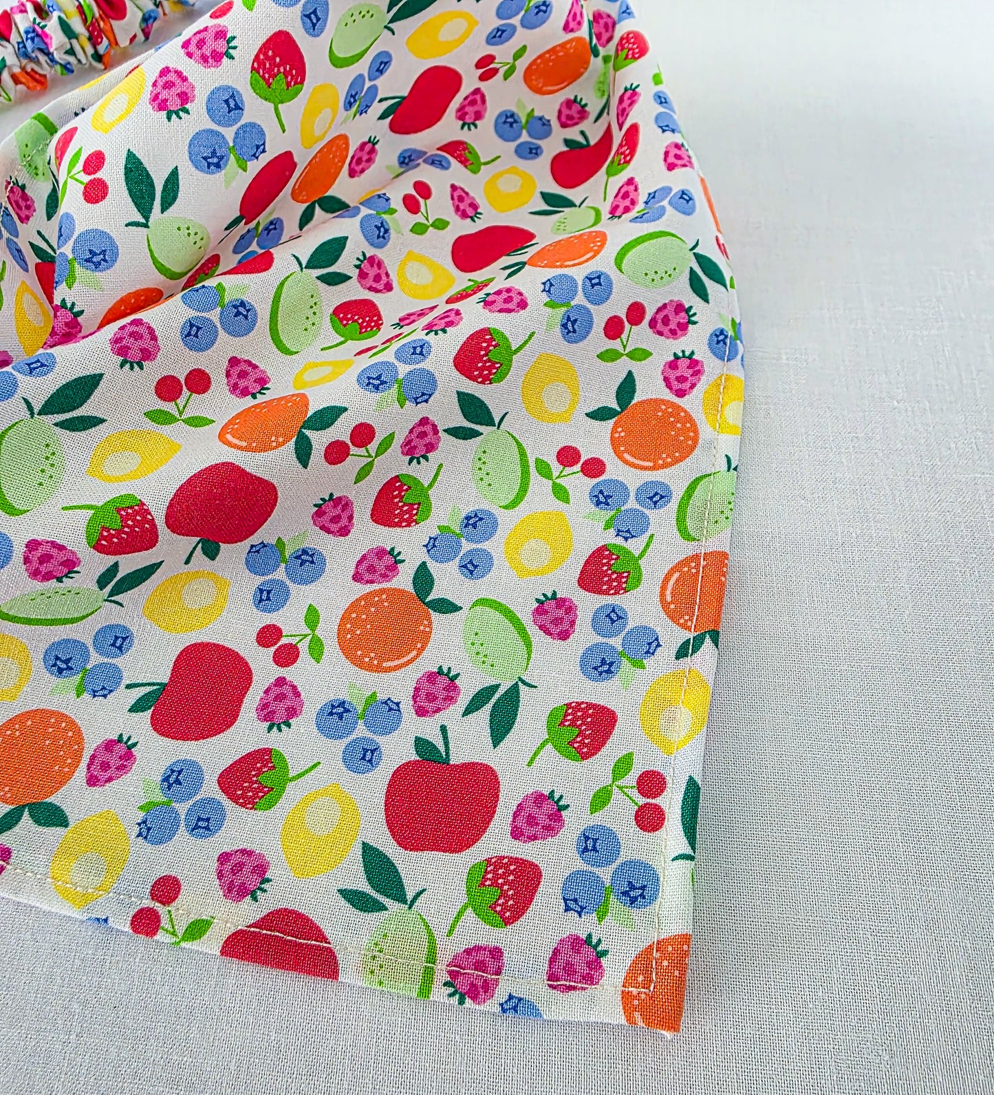 A close-up of a white fabric with a colorful fruit and berry print, laid out on a light gray textured surface.