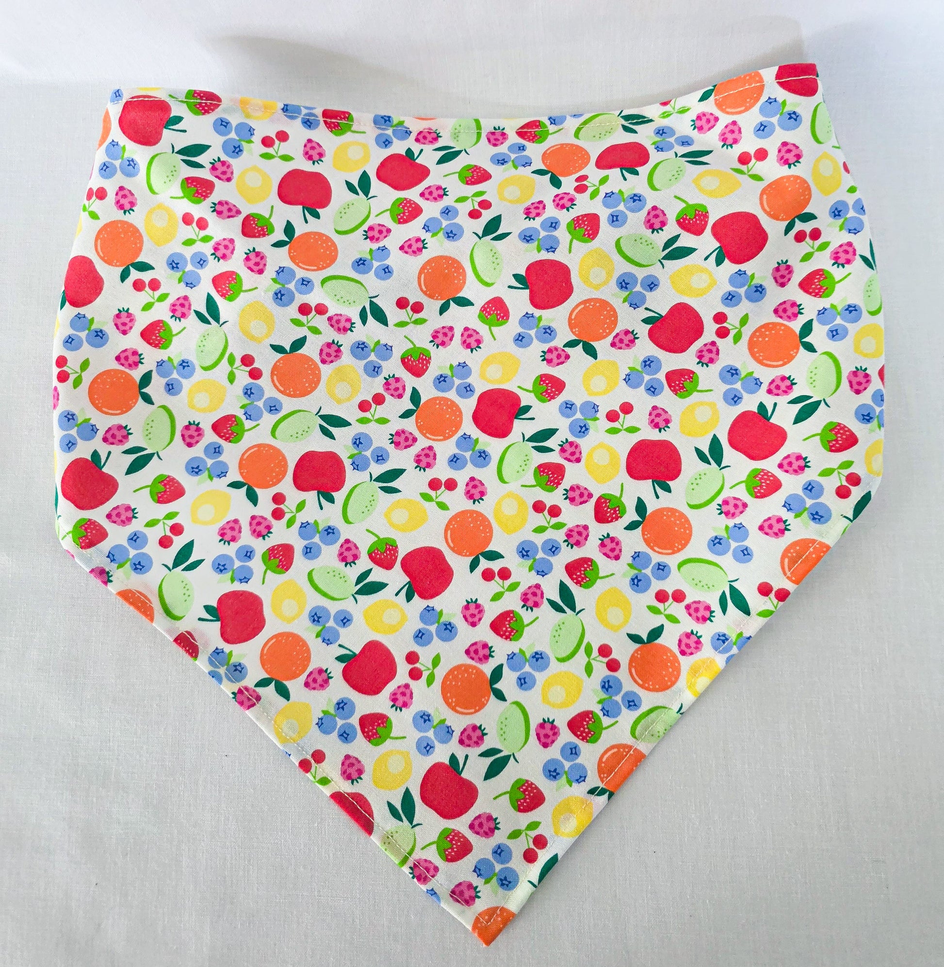 A triangular bandana-style hair bandana featuring a colorful fruit and berry print on a white background, laid flat on a light fabric surface.