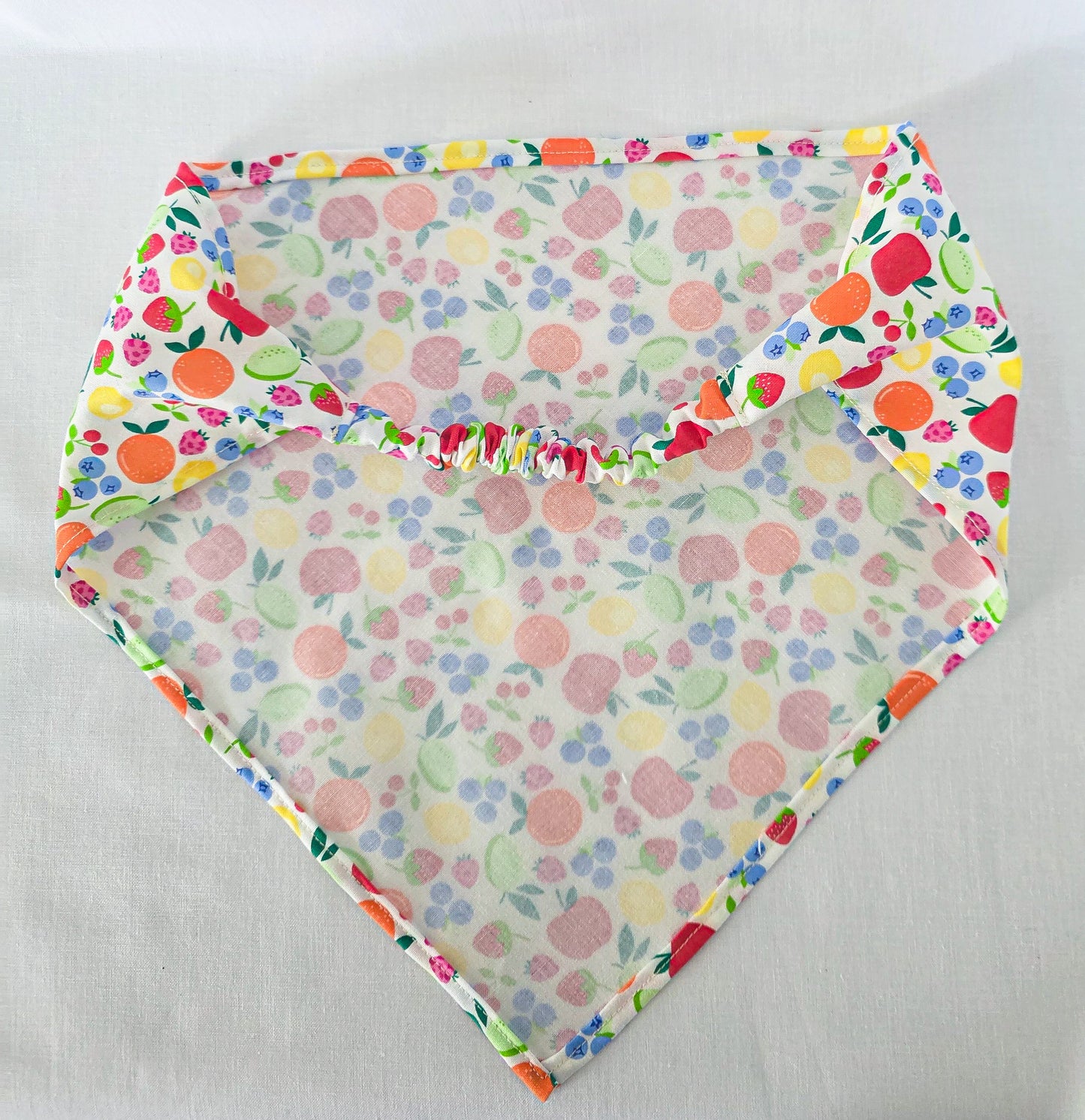 A triangular hair bandana laid flat on a light fabric background. The bandana has a colorful fruit and berry print on a white base.