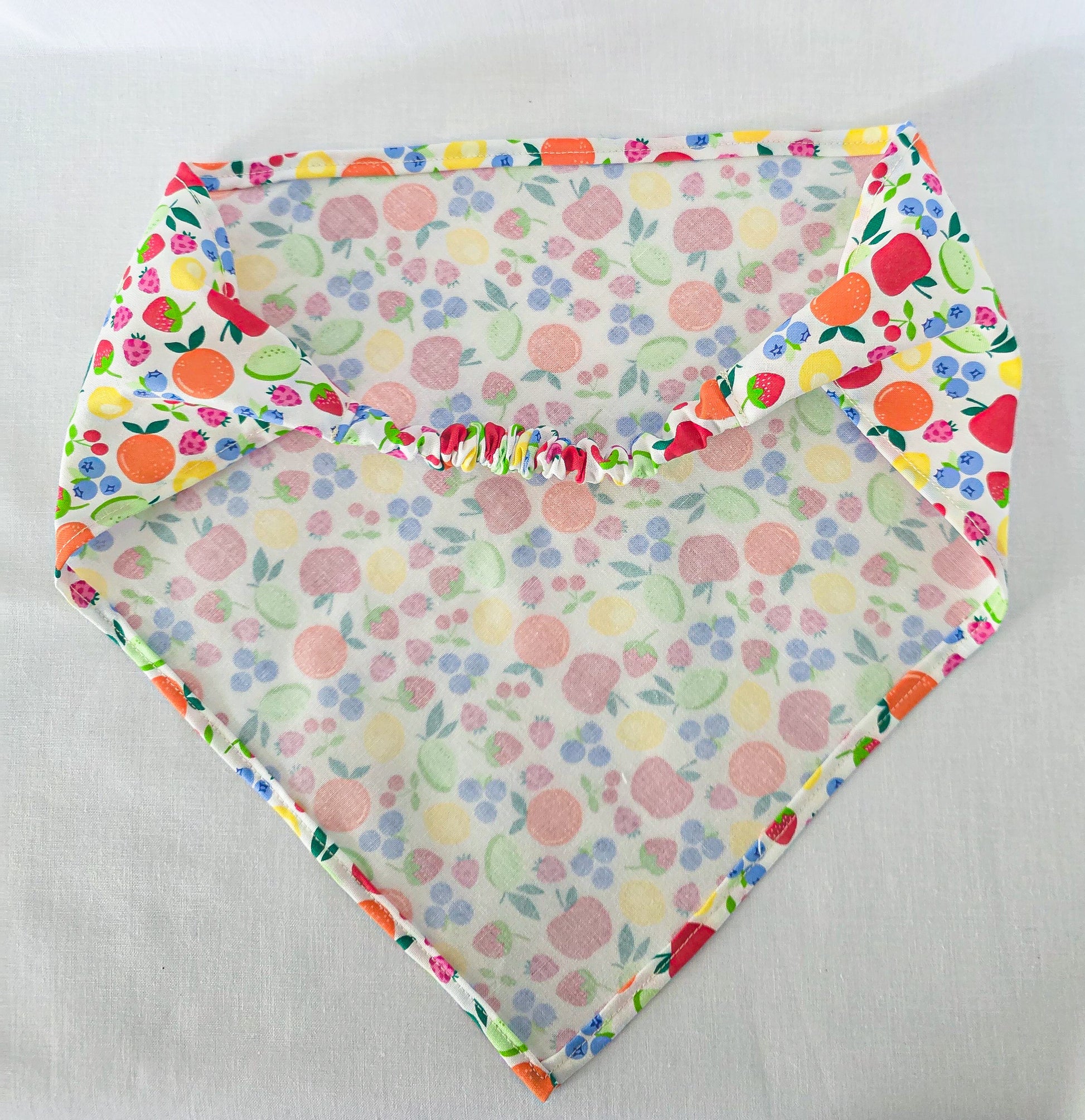 A triangular hair bandana laid flat on a light fabric background. The bandana has a colorful fruit and berry print on a white base.