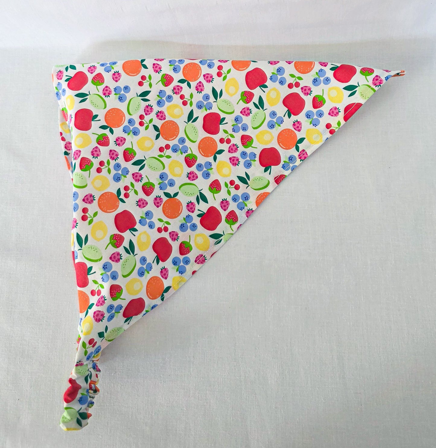 A triangular bandana  featuring a colorful fruit and berry print on a white background, laid flat on a light fabric surface.