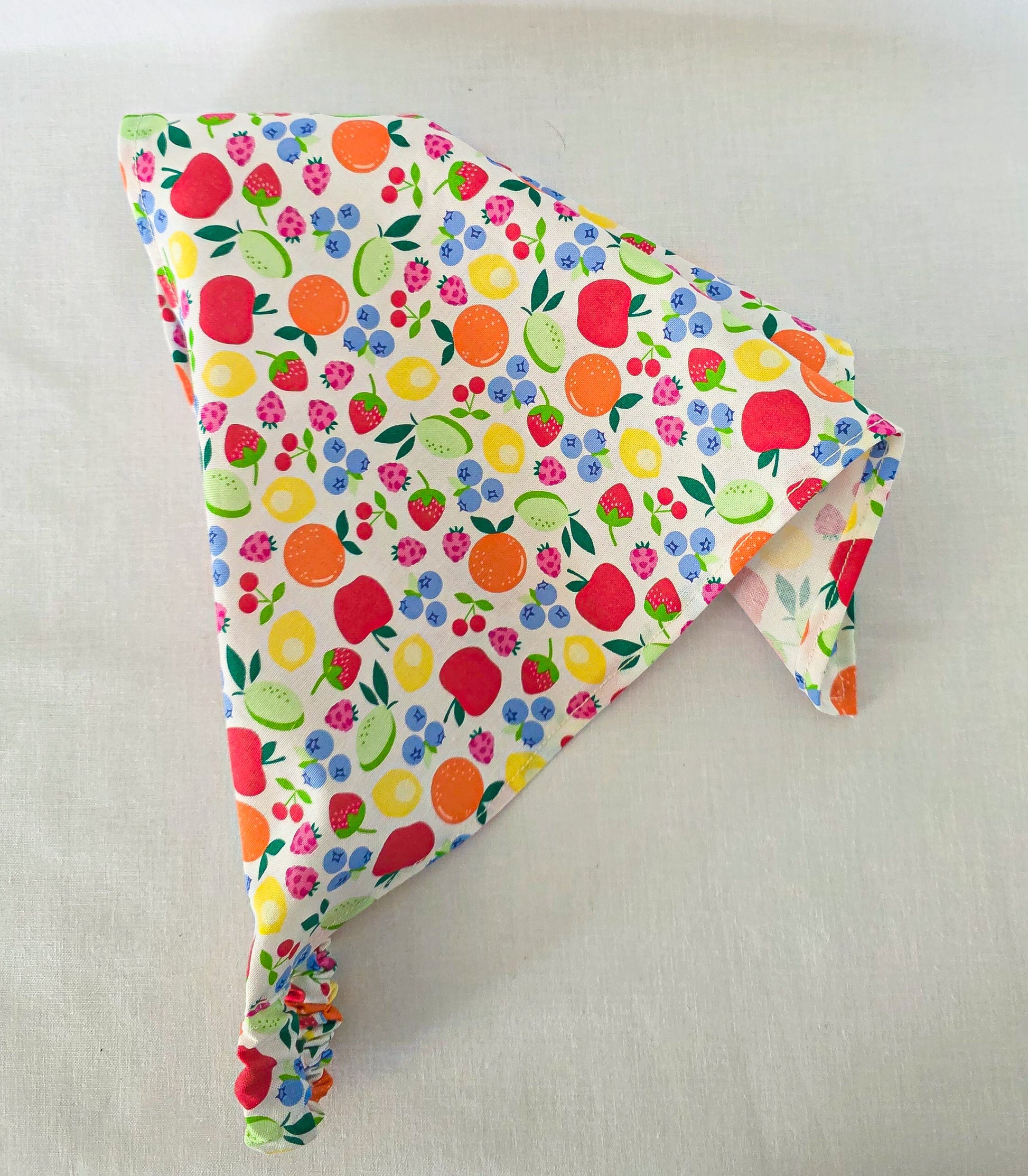 A triangular hair bandana made of white fabric with a colorful fruit print, laid flat on a light fabric background.