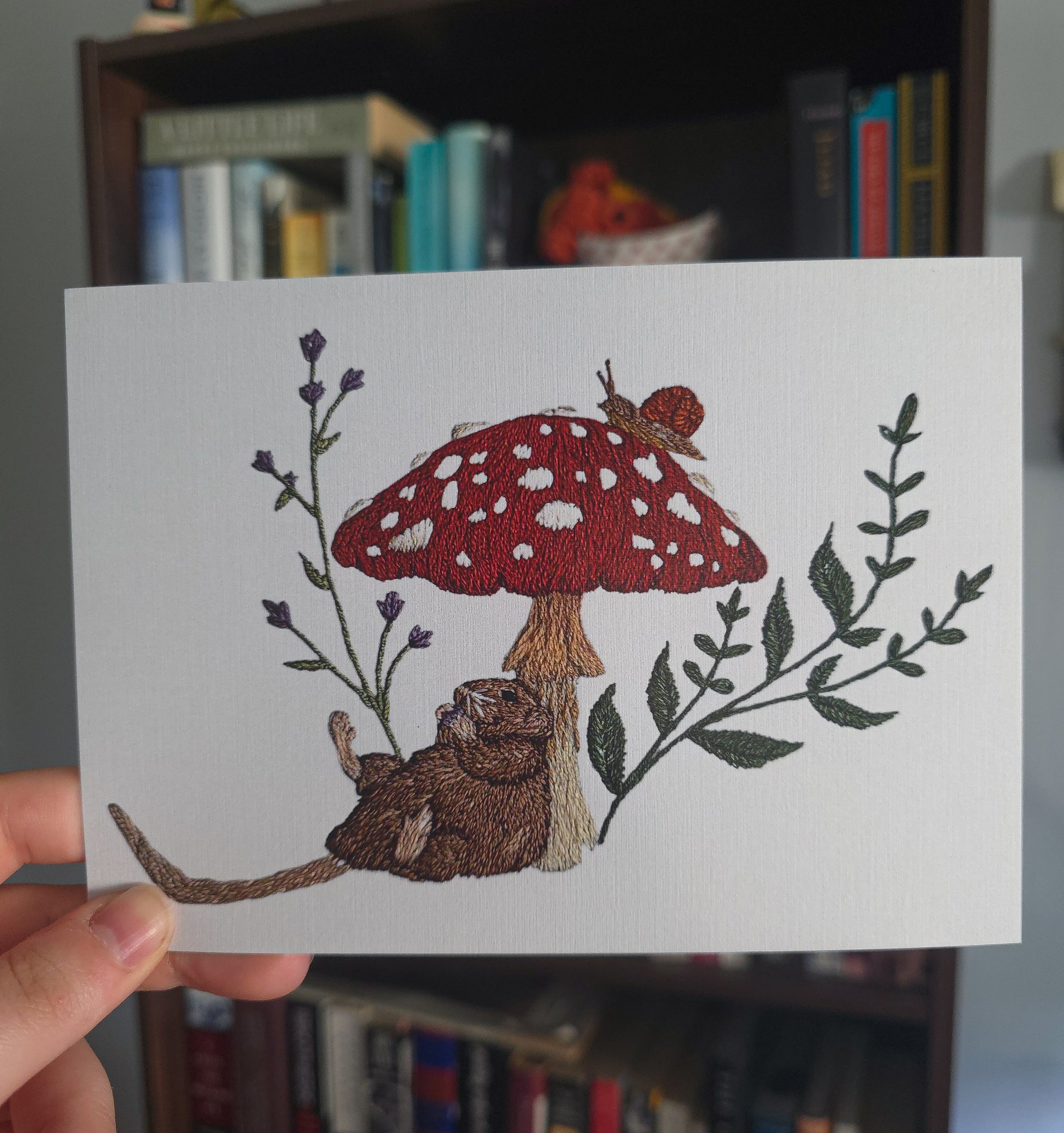 A hand holds a white embroidered art print featuring a red mushroom with white spots, a brown rat, green leaves, and small purple flowers, with a blurred bookshelf in the background.
