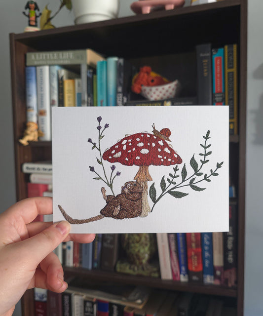 A hand holds a white art print featuring a whimsical embroidered-style illustration of a small brown rat under a red-and-white spotted mushroom, with green leaves and purple flowers. The background shows a dark wooden bookshelf filled with books.