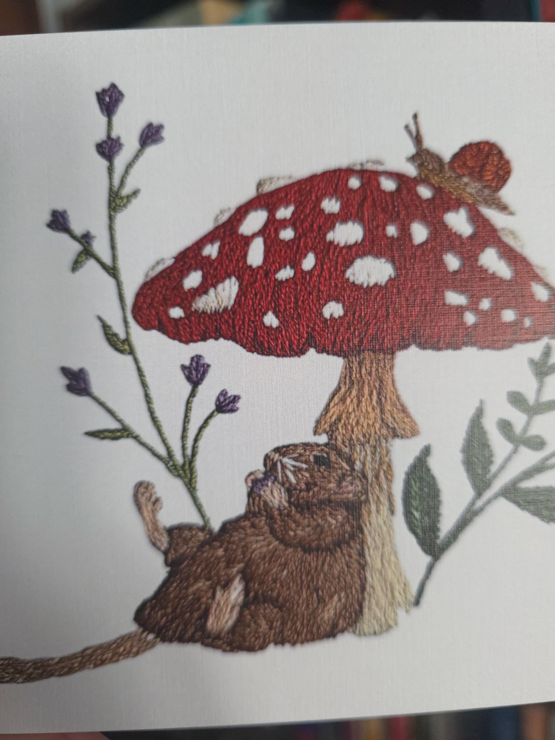 a print of and embroidered woodland design featuring a brown rat under a red and white spotted mushroom, with green leaves and purple flowers on a white background.
