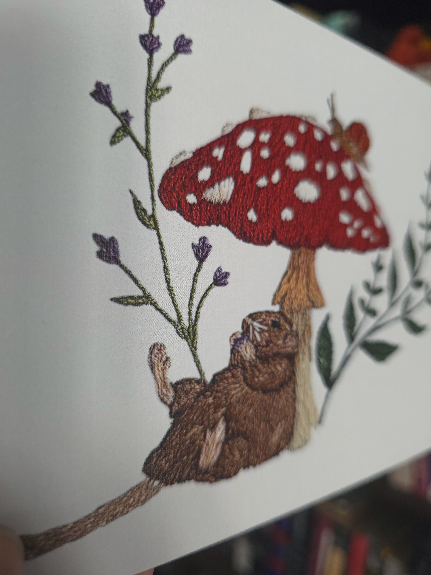 A close-up of a white embroidered artwork featuring a brown rat sitting beneath a red-and-white spotted mushroom, with purple flowers and green leaves around it.