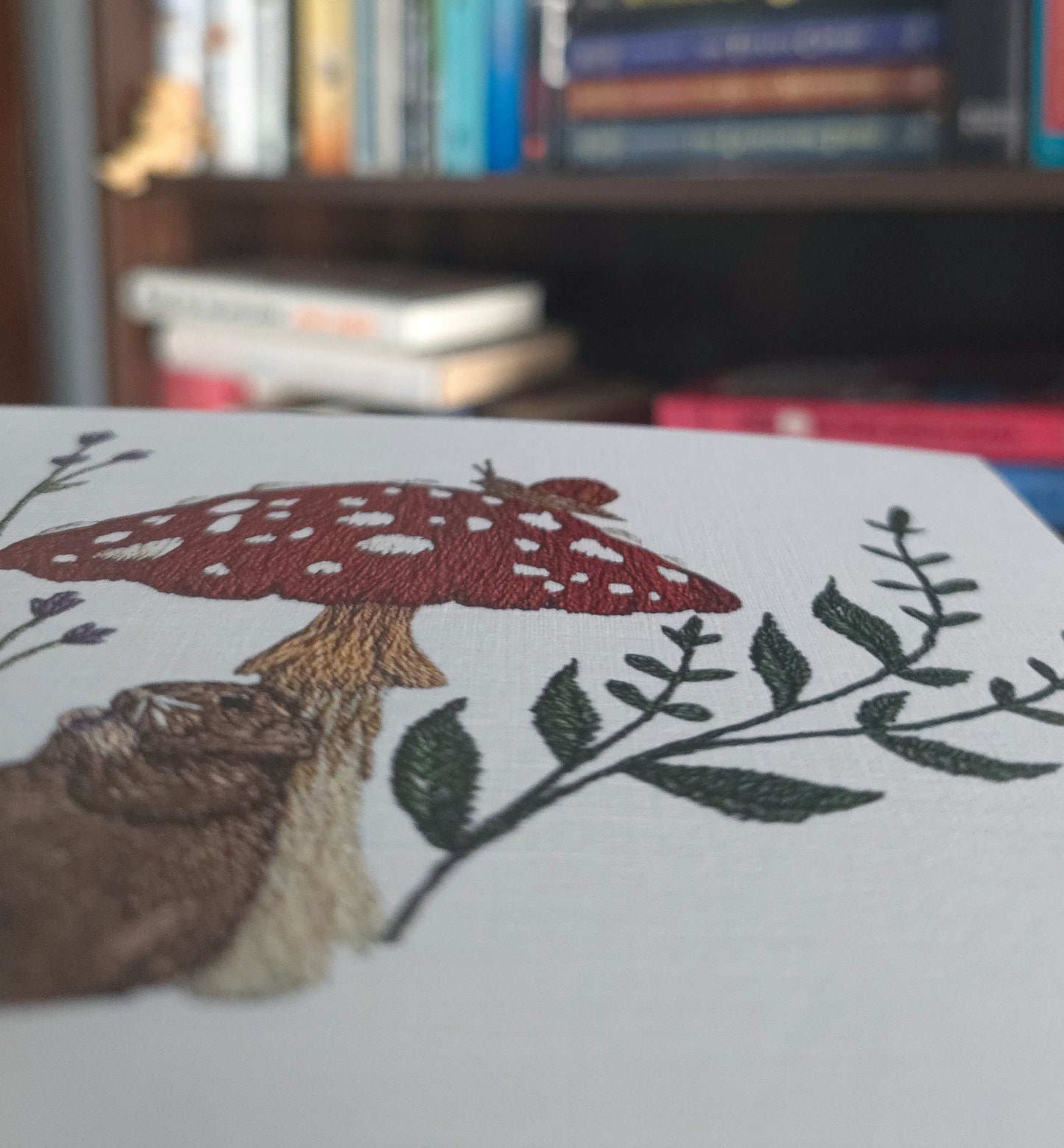 A close-up of a white embroidered print featuring a red and white spotted mushroom, a brown rat, and green leafy branches, with a blurred bookshelf in the background.