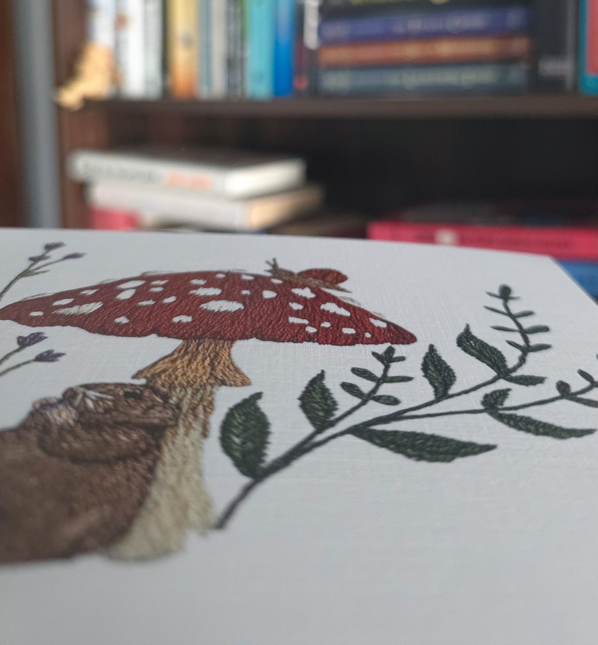 A close-up of a white embroidered print featuring a red and white spotted mushroom, a brown rat, and green leafy branches, with a blurred bookshelf in the background.