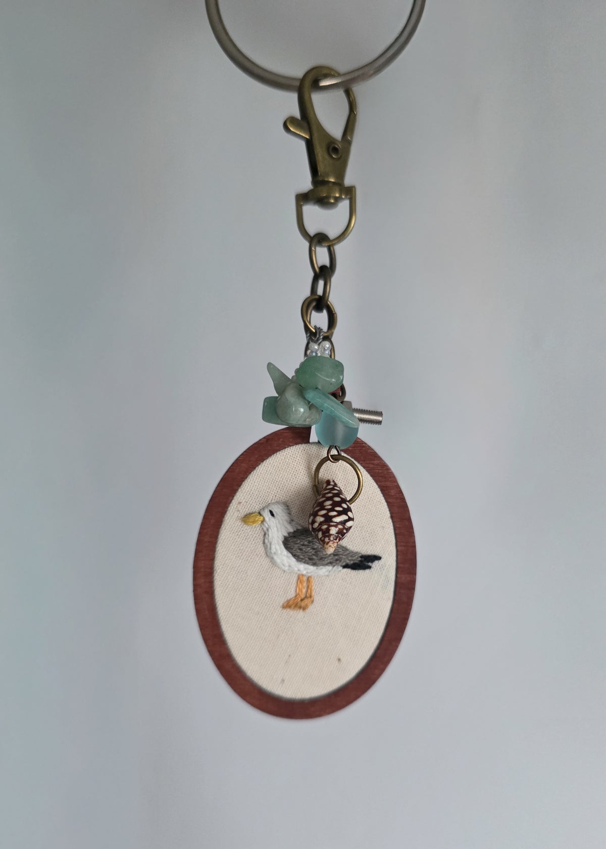 A handmade keychain featuring a small oval wooden frame with a cream background , decorated with a detailed seagull figure. The keychain includes a brass clasp, a green stone bead, and a small shell charm, set against a plain light gray background.