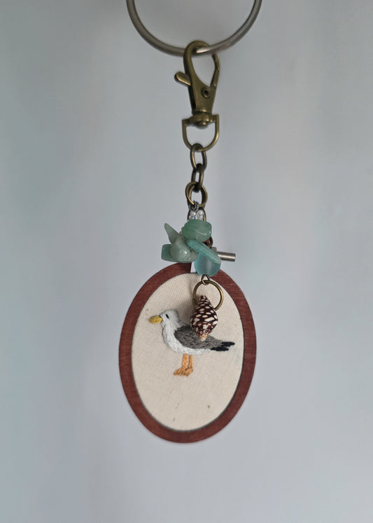 A handmade keychain featuring a small oval wooden frame with a cream background , decorated with a detailed seagull figure. The keychain includes a brass clasp, a green stone bead, and a small shell charm, set against a plain light gray background.