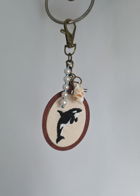 A handmade keychain featuring an oval wooden pendant with a black and white orca whale, decorated with white faux pearls, a small conch shell, and metallic beads, hanging from a brass key ring.