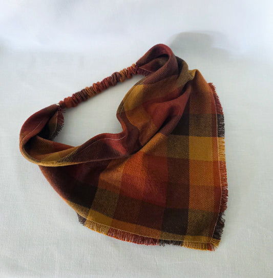 A plaid bandana in warm autumn colors, featuring a fringed edge and elasticized strap, laid out on a white fabric background.
