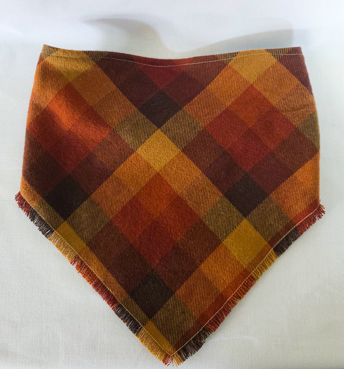 A triangular bandana with a warm autumn plaid pattern in orange, brown, and yellow, featuring fringed edges, laid flat on a white background.