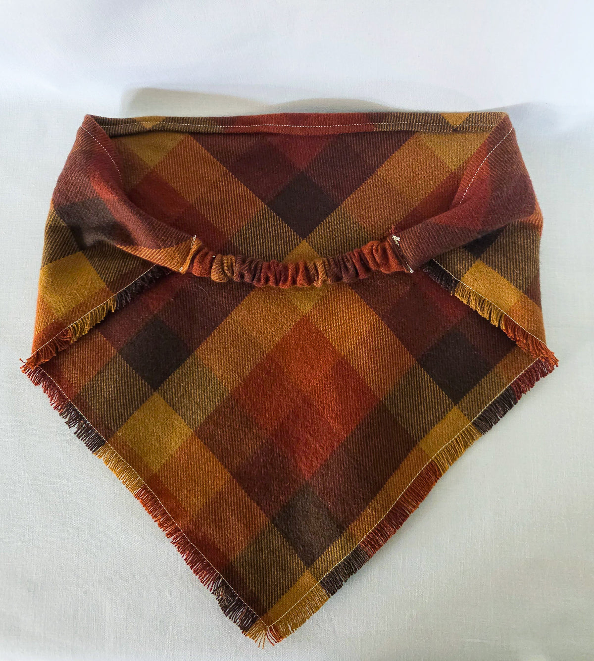A plaid bandana featuring a warm autumn color palette of rust orange, mustard yellow, brown, and dark red. The top band is fringed, and the bottom corners are also frayed.
